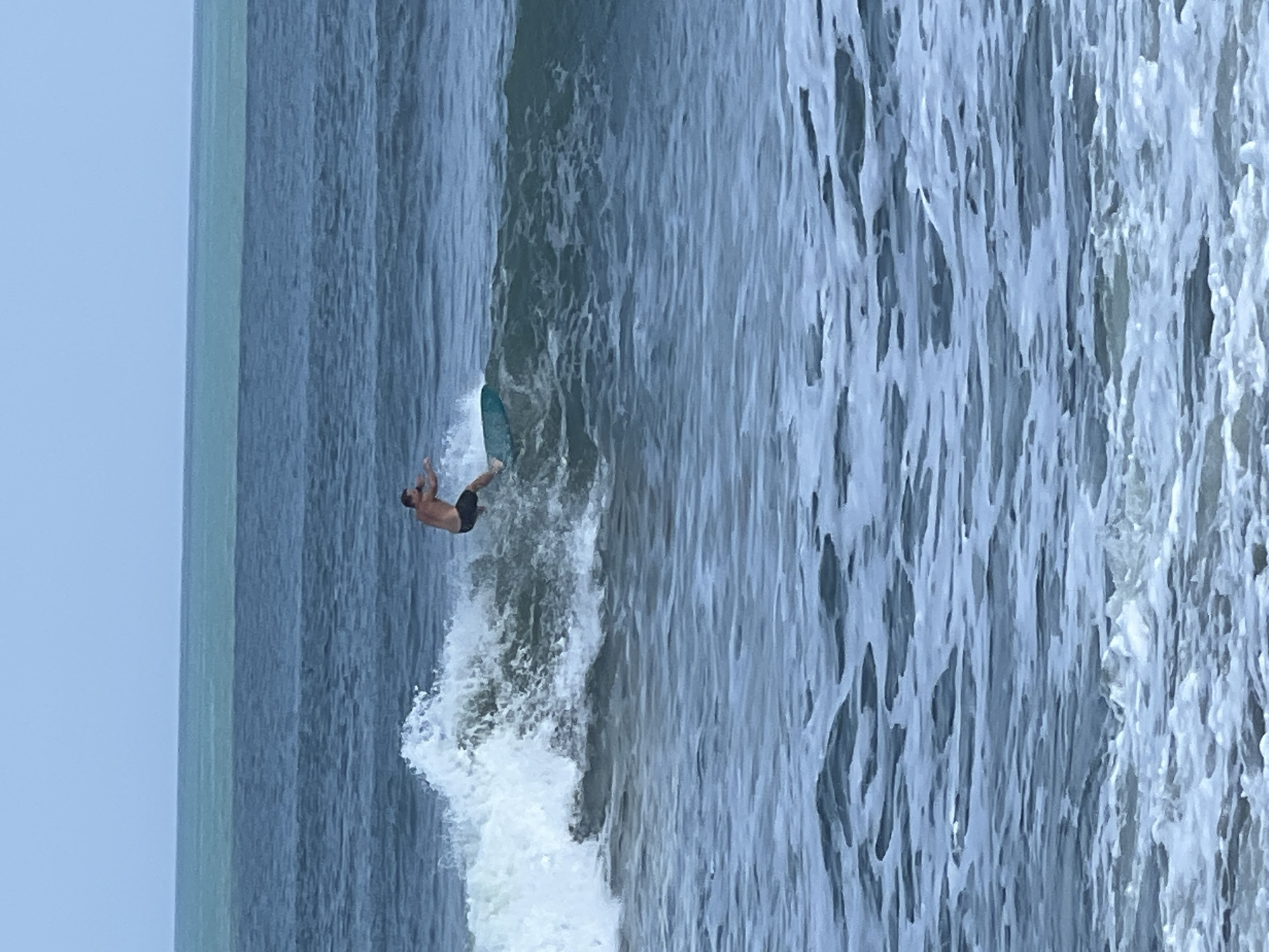 A surfer is riding a wave in the ocean. The surfer is a man wearing black shorts. He is on a teal surfboard and is in the middle of the wave, leaning to his right. The water is blue with white foam where the wave is breaking. The sky is overcast and a pale blue. The ocean has multiple layers of blue water, with a lighter turquoise band in the distance. This scene is likely Kuta, Indonesia, a popular surfing destination known for its consistent waves and beaches.