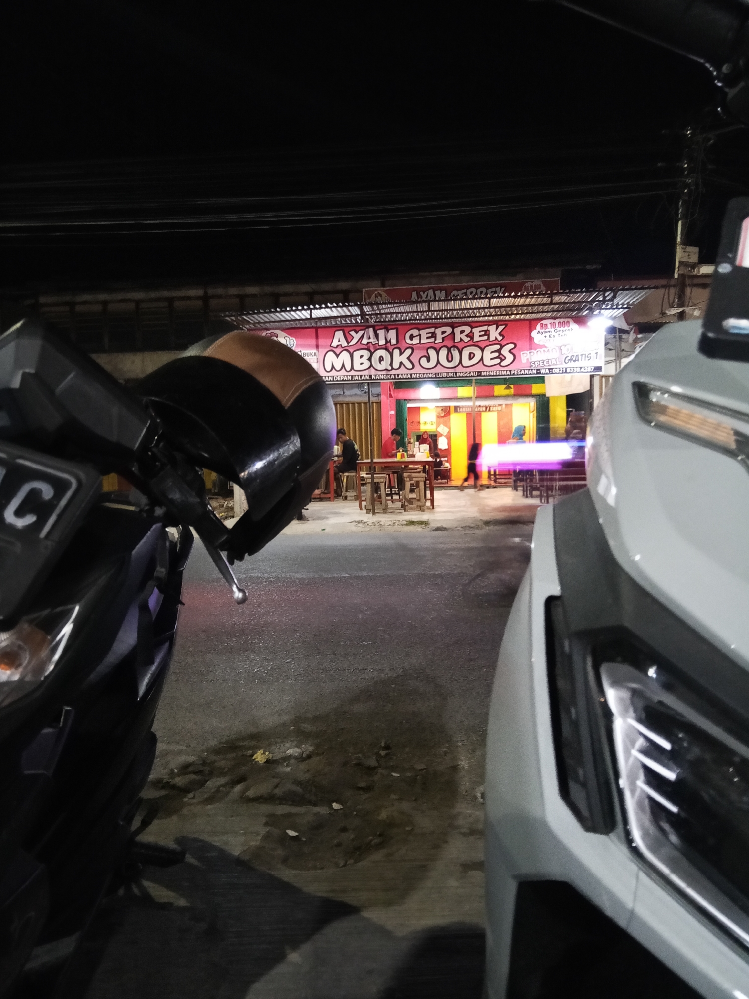 A low-angle, night-time shot captures the front of two parked motorcycles. The motorcycle on the left is dark-colored with a partially visible license plate and a black and brown helmet hanging from its handlebars. The motorcycle on the right is light gray and takes up the right third of the frame.
In the background, a brightly lit food stall with a red banner announces "AYAM GEPREK MBOK JUDES" and offers a "PROMO 10.000 SPECIAL GRATIS 1". The stall features yellow and green walls, with tables and chairs set up outside. Two individuals are visible inside the stall, one standing and the other seated. Another person is walking in front of the stall. The ground in front of the stall appears to be a paved road with some debris. The overall scene suggests a busy street food environment in the city of Lubuklinggau, Indonesia, at night.