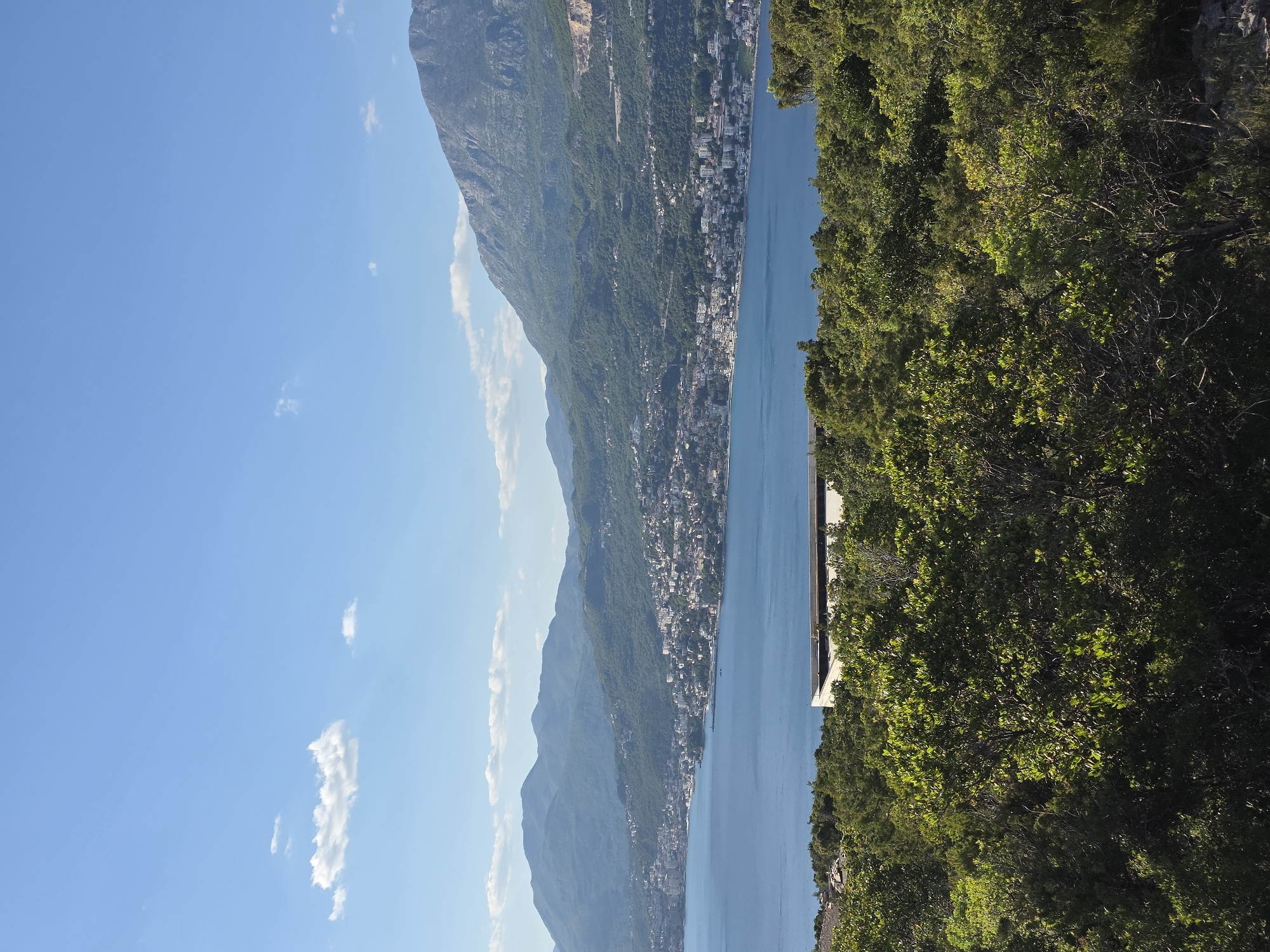 This landscape image presents a scenic coastal view under a bright, clear blue sky dotted with a few wispy white clouds. On the left side, a majestic mountain range dominates the horizon, its slopes richly covered in dense green vegetation and some visible rocky outcrops. At the base of these mountains, along the curving shoreline, a settlement of numerous white and light-colored buildings stretches, indicating a vibrant coastal town or village.
A calm, deep blue body of water, likely a bay or a section of the sea, occupies the central part of the image, reflecting the bright sunlight. The water appears tranquil, with no visible boats or activity.
The right foreground is filled with dense, lush green trees and shrubbery, suggesting the photograph was taken from an elevated vantage point within a forested or heavily vegetated area. Partially visible within these trees, on the right side overlooking the water, is a small, modern-looking building with a flat, light-colored roof.
The scene depicts a sunny day, likely morning or early afternoon, with excellent visibility. No people, specific activities, or discernible text are visible in the photograph. The setting is characteristic of a mountainous coastal region, consistent with the provided context of Luštica, Montenegro.