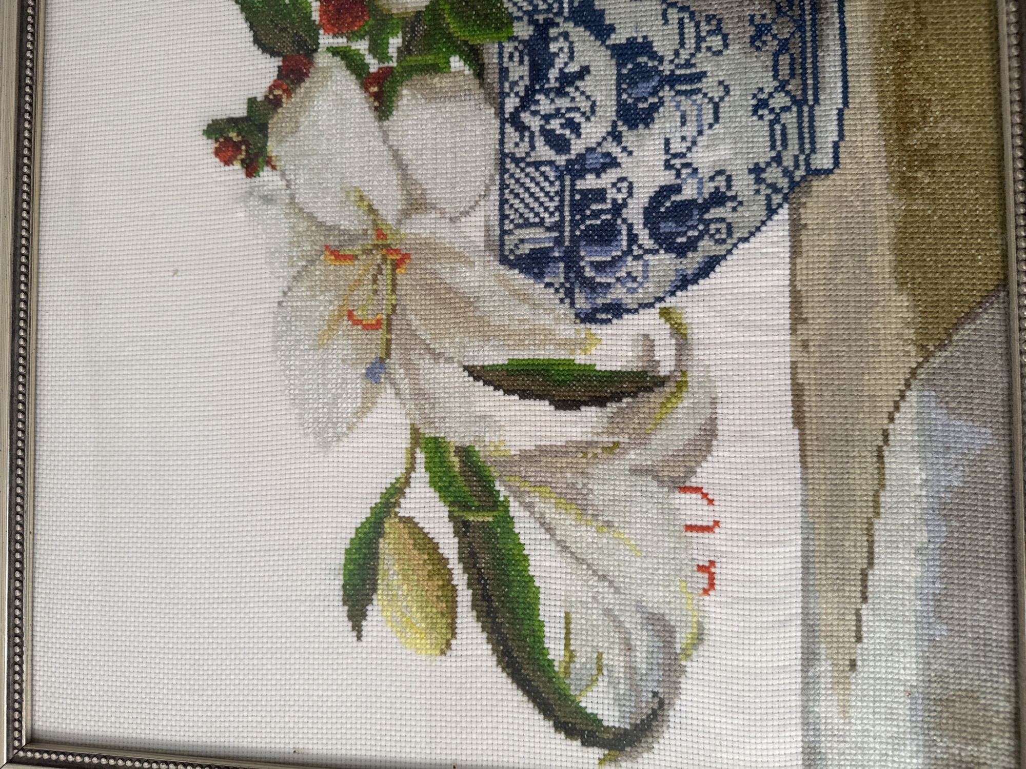 A cross-stitch artwork depicting a still life of lilies in a blue and white porcelain vase is displayed within a silver frame. The artwork features white lilies with yellow and orange accents in their centers, green leaves, and hints of red berries and green foliage. The vase has a traditional blue and white floral pattern. The background is a plain white canvas. The artwork is placed on a surface that appears to be a table or shelf with a textured, neutral-toned fabric covering it. The overall scene is static and artistic, with no signs of people or activity. There is no indication of the city of Bishkek, Kyrgyzstan, in the image.