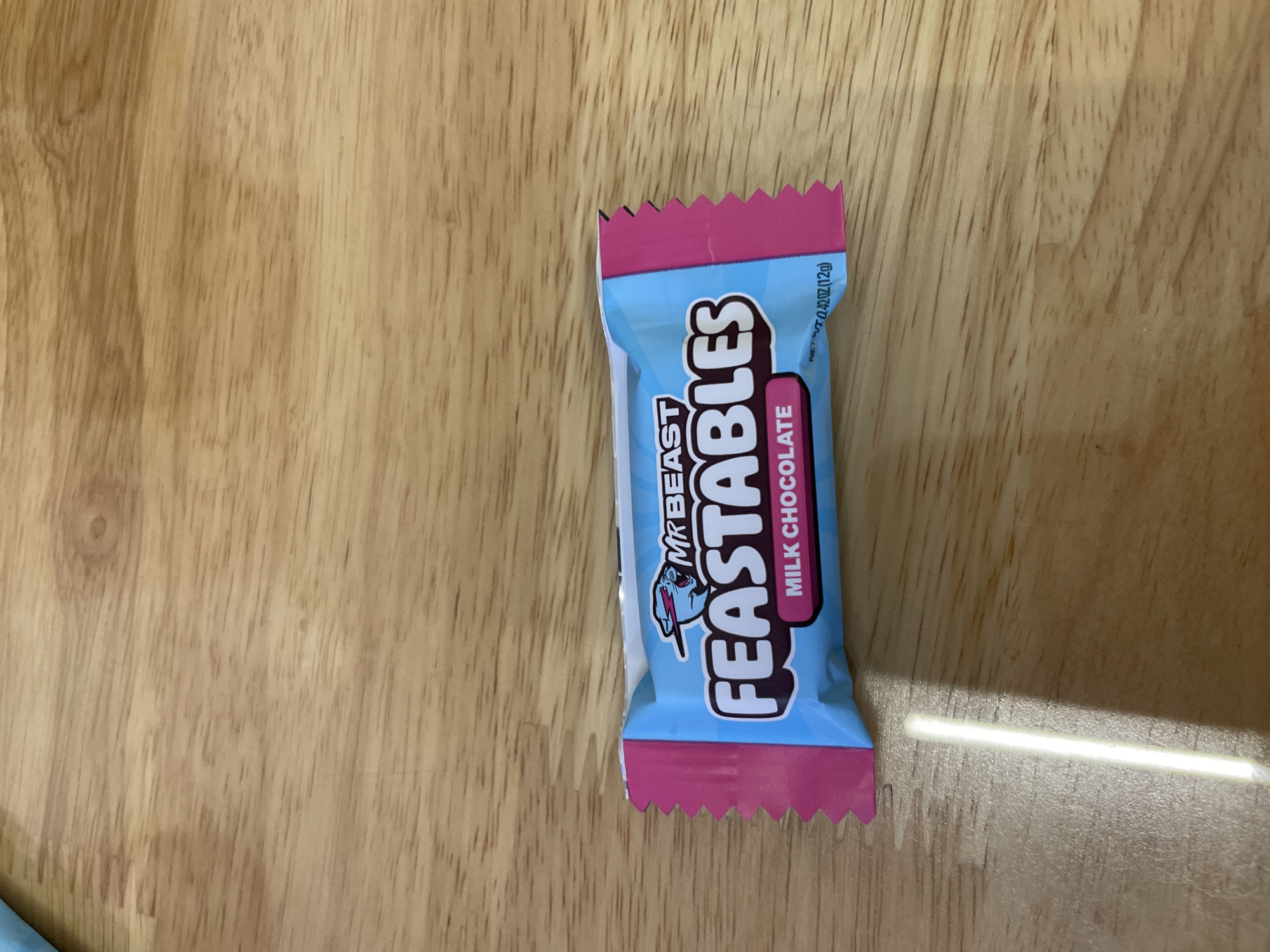 A single MrBeast Feastables milk chocolate bar is positioned on a wooden surface. The bar is small and rectangular, sealed in a light blue wrapper with pink accents and the "Feastables" logo prominently displayed. The wooden surface has a natural grain pattern. The lighting is bright, creating a distinct shadow to the right of the candy bar. The image doesn't suggest a location in Bắc Nha Trang, Vietnam.