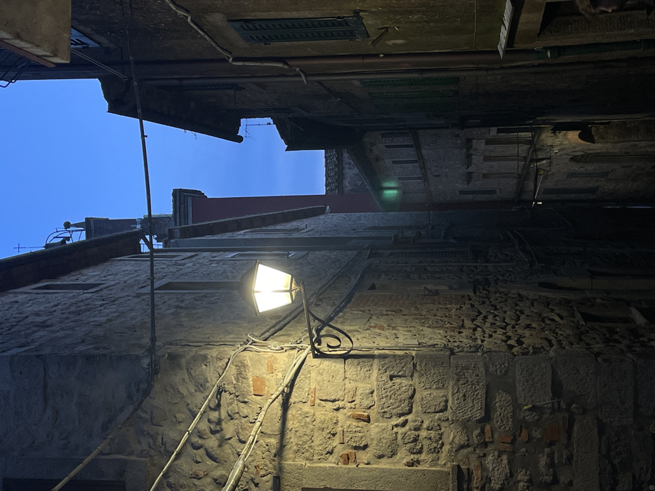 An image captures a narrow, illuminated alleyway in Castel del Piano, Italy, at twilight. The scene is dominated by two ancient-looking buildings constructed from rough-hewn stone blocks, defining the confined space. On the left, a building with visible reddish brickwork interspersed among the stone features multiple grey electrical conduits and wires running horizontally and vertically along its facade. An ornate black metal streetlamp, featuring a square lantern, extends from this wall on a scrolled bracket, emitting a warm yellow-white light that illuminates the immediate foreground and casts long shadows.
Opposite, on the right, a darker stone building displays several closed, dark green wooden shutters on its windows. Deeper into the alley, additional stone buildings become visible, one exhibiting a faint green glow from an interior window. The ground surface of the alleyway is mostly obscured by shadow. Above the structures, a clear twilight sky with a deep blue hue is visible, bordered by the rooftops. A reddish building section is present in the mid-distance, and above it, a small cross or finial is silhouetted against the sky, suggesting a religious or historical structure. In the lower right corner, the top of a human head and part of a shoulder are visible, partially obscured by shadow, indicating a person standing near the vantage point of the camera. No explicit actions or interactions are depicted.