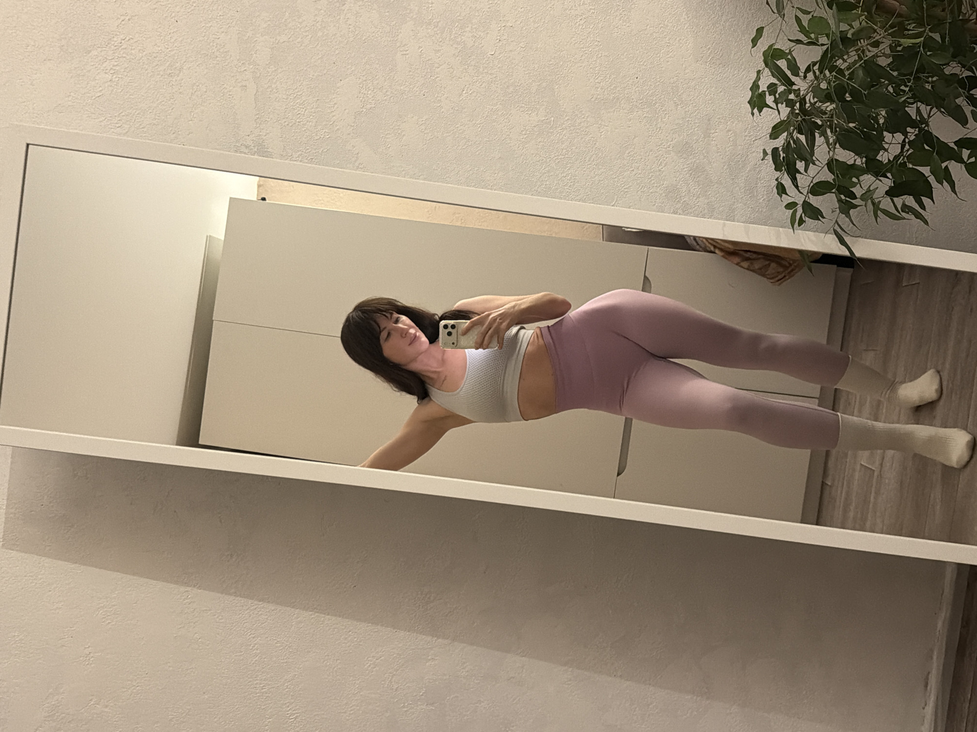 A woman is depicted taking a mirror selfie in an indoor setting, likely in Perm, Russia. She is lying or leaning on her side, posing for the camera. She has dark hair with bangs and is dressed in a light grey sports bra, light purple leggings, and white socks. Her expression is calm, with a slight smile. She holds a light-colored smartphone, possibly an iPhone, in her right hand, using it to capture her reflection.
The full-length mirror, framed in white, occupies most of the frame. In the reflection, a large, light-colored wardrobe or cabinet with multiple doors is visible behind the woman. To the upper right of the reflection, a potted plant with numerous green leaves adds a natural element to the room. The floor appears to be made of light-toned wood or laminate. The wall against which the mirror is placed has a textured, off-white surface. The lighting in the room appears to be artificial, indicating an indoor environment, possibly during the evening or night. There is no visible text in the image.