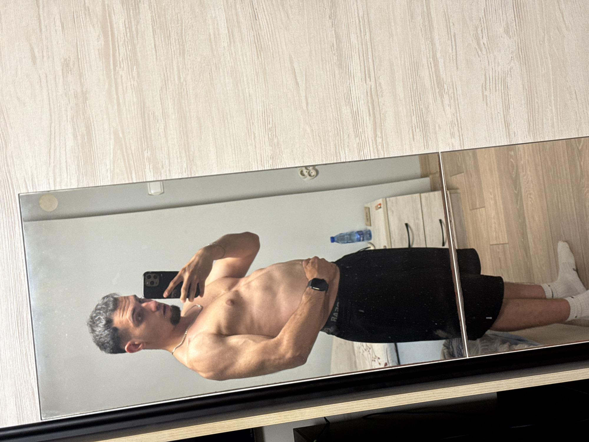 A man is taking a mirror selfie in what appears to be a bedroom. He is shirtless, wearing black shorts, white socks, and a watch on his left wrist. He has short, dark hair with a fade haircut and a goatee. In the background, a nightstand with a bottle of water and a drawer is visible. The floor is wooden. The image is framed by a mirror and a wooden wardrobe. The location is not specified in the image.