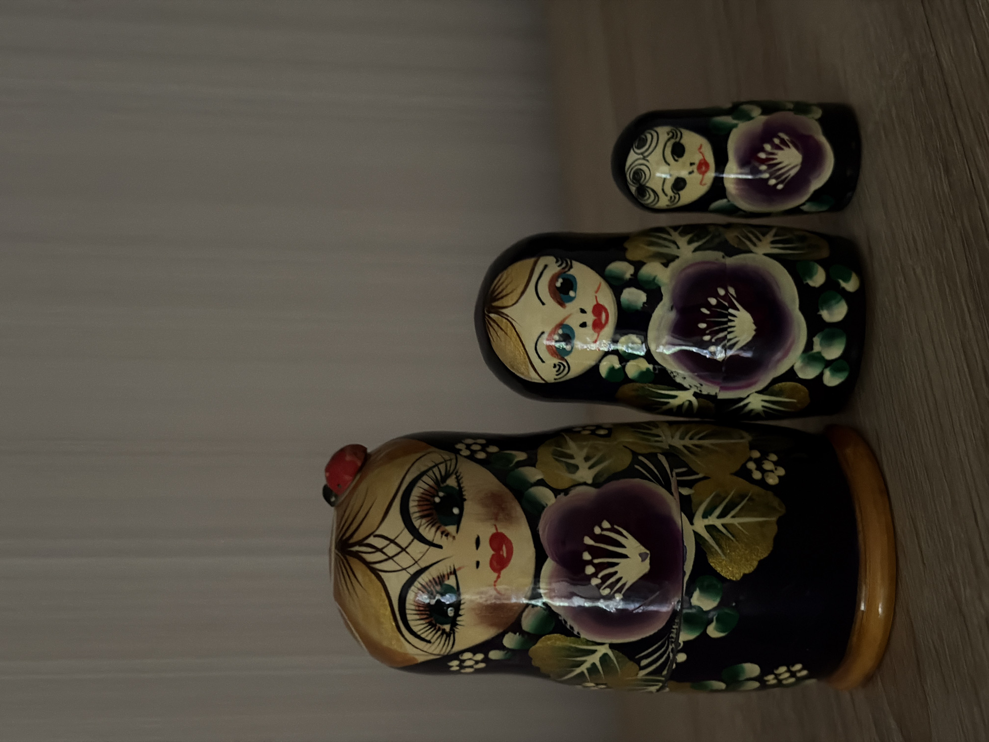 Three Matryoshka dolls are arranged on a wooden surface, with a light-colored, vertically paneled wall in the background. The largest doll is on the left, a medium-sized doll is in the center, and the smallest doll is on the right. All dolls are painted with similar designs featuring faces, flowers, and decorative patterns. The largest doll has a red ladybug accessory on its head. The lighting is dim, casting soft shadows. The image does not provide any information about the city of Cheltuitori, Moldova.