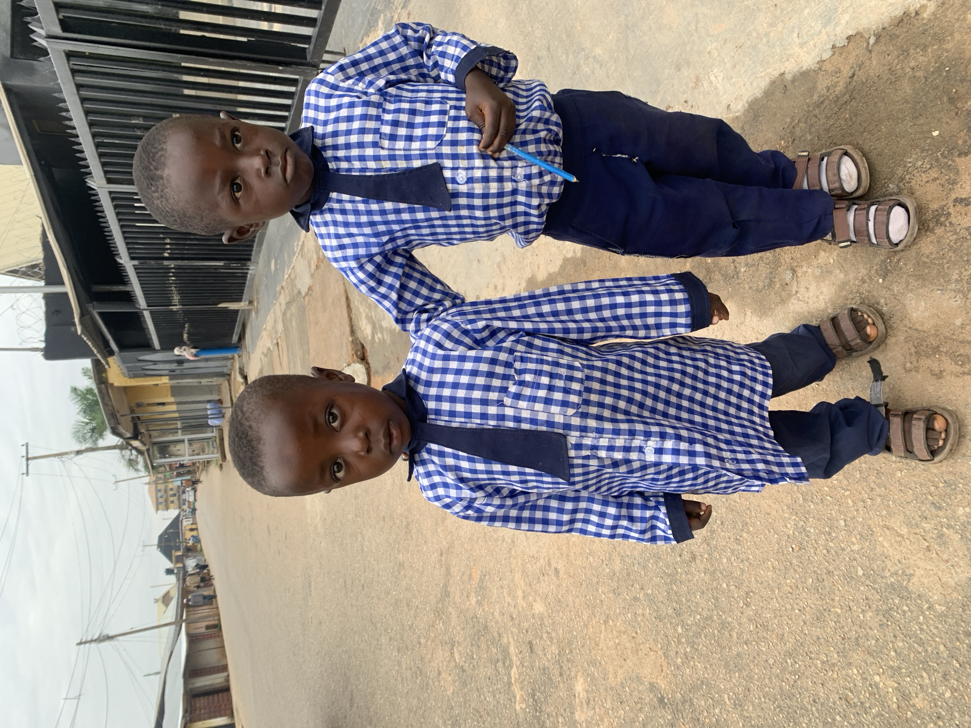 Two young boys in blue and white checkered school uniforms with navy blue ties and pants stand on a paved street in Jos, Nigeria. The boy on the left is closer to the camera and looks directly at the viewer. The boy on the right stands slightly behind and to the right of the first boy, holding a blue pen in his right hand and looking forward. Both boys are wearing brown sandals. The street is lined with buildings and a black metal fence. The sky is overcast.