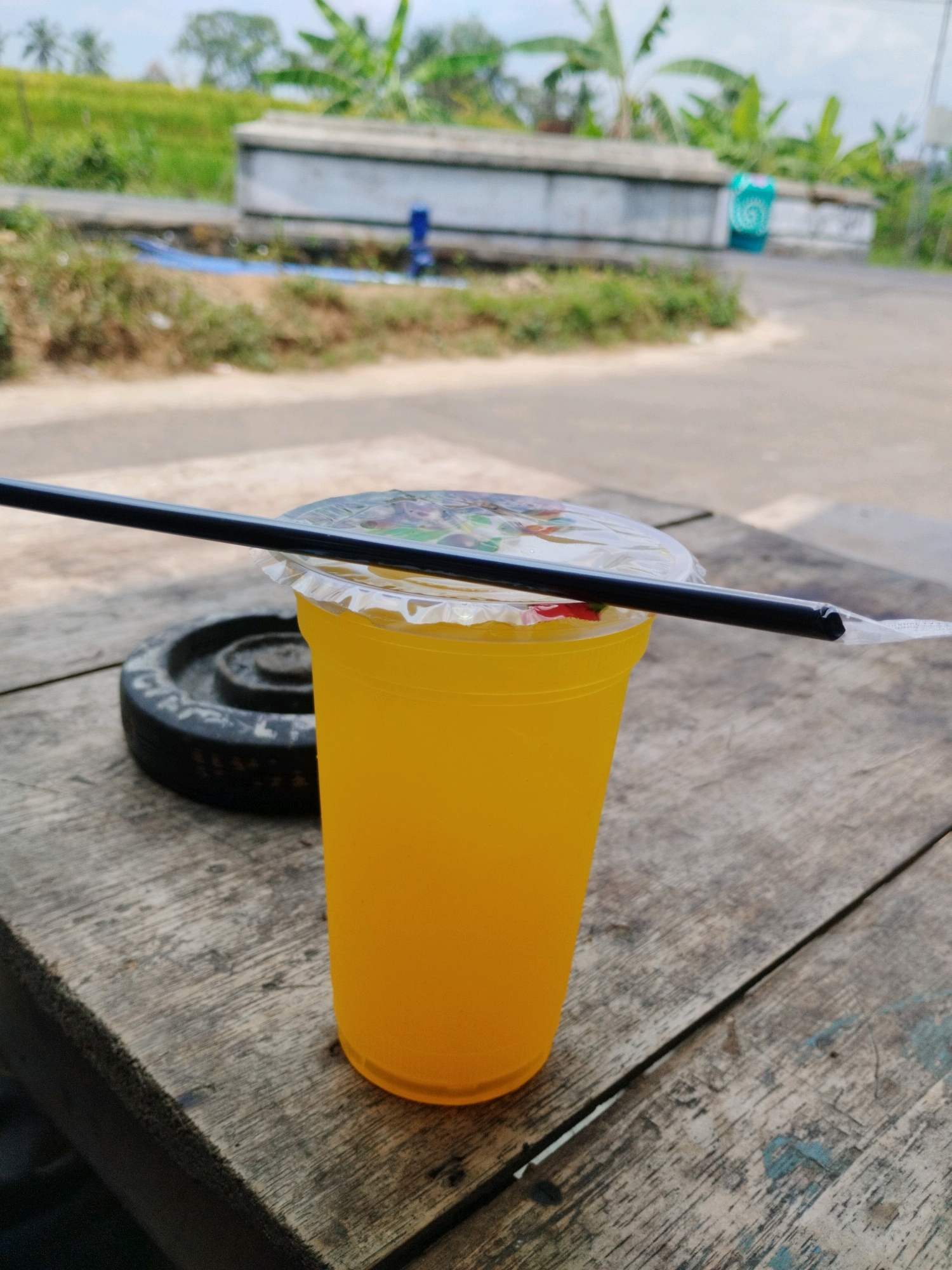 A plastic cup of orange-colored liquid with a clear lid and a black straw is on a weathered wooden table. To the left of the cup is a black circular object, possibly a weight. In the background, there is a paved road, a grassy embankment, and lush green foliage including banana trees. In the distance, there are some structures, including a concrete wall. The sky is overcast. The scene appears to be outdoors in a rural or semi-rural area of Pakuncen, Indonesia.