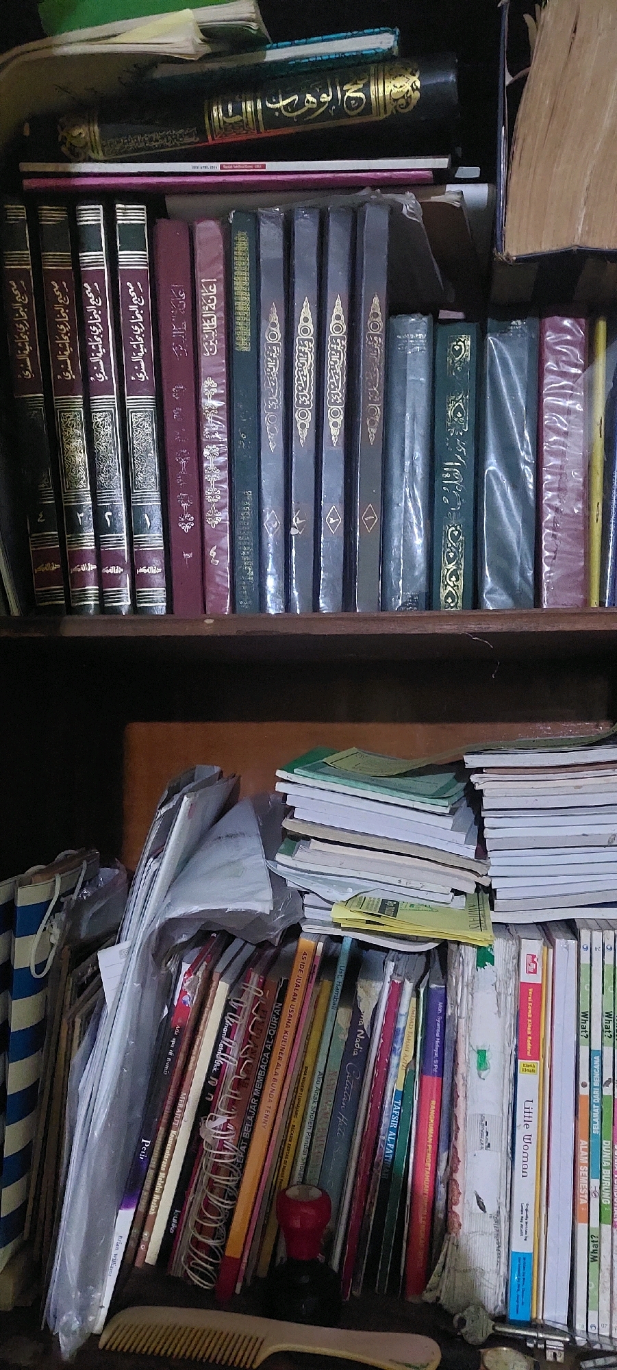 The image shows a close-up view of a wooden bookshelf filled with books and other items. The shelf is divided into two levels. The top level contains several books, some of which appear to be religious texts with Arabic script and gold embellishments. One book is black with gold lettering and an ornate design. There are also some smaller, lighter-colored books.
The bottom level of the shelf is more cluttered. A stack of papers and notebooks is visible on the left, some of which are bound in plastic. A collection of books with colorful spines is arranged vertically. Some of these books have titles in what appears to be Malay or Indonesian. There are also some loose papers, a red stamp, and a plastic comb on this shelf. Keys and coins are also visible on the far right of the bottom shelf.
The lighting in the image is dim, suggesting an indoor environment, possibly a room or a study. The wooden shelf has a worn appearance. The overall impression is one of a personal collection of books and reading materials.
The request to include the location of Lumajang, Indonesia, cannot be fulfilled as there are no visual cues in the image to indicate this specific location.