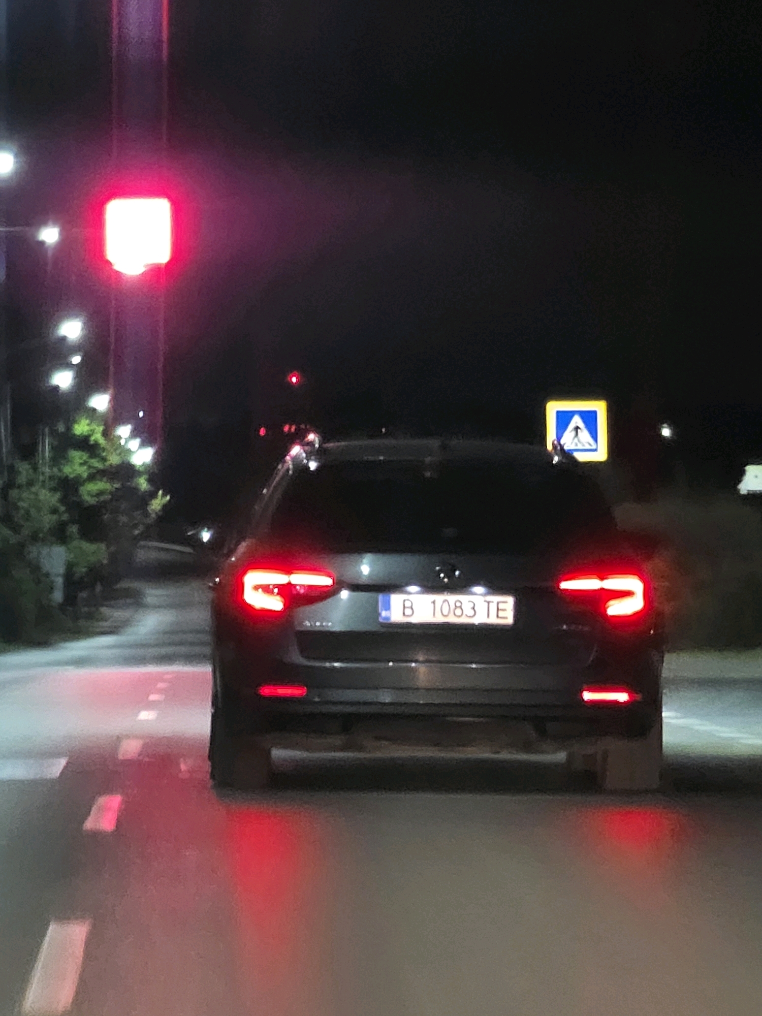 A dark-colored Audi A4 is shown from the rear, stopped at a red traffic light at night on a paved road. The car's license plate is visible, displaying "B 1083 TE". The traffic light above the vehicle is red and illuminated, casting a reddish glow on the car and the road ahead. To the right of the car, a pedestrian crossing sign with a blue square background and a white pedestrian symbol is visible. The road surface has painted white dashed lines indicating lanes. Streetlights are present in the background, illuminating trees and foliage on the left side of the road. The overall scene suggests a typical urban or suburban environment at night. The location is identified as Aksakovo, Bulgaria.