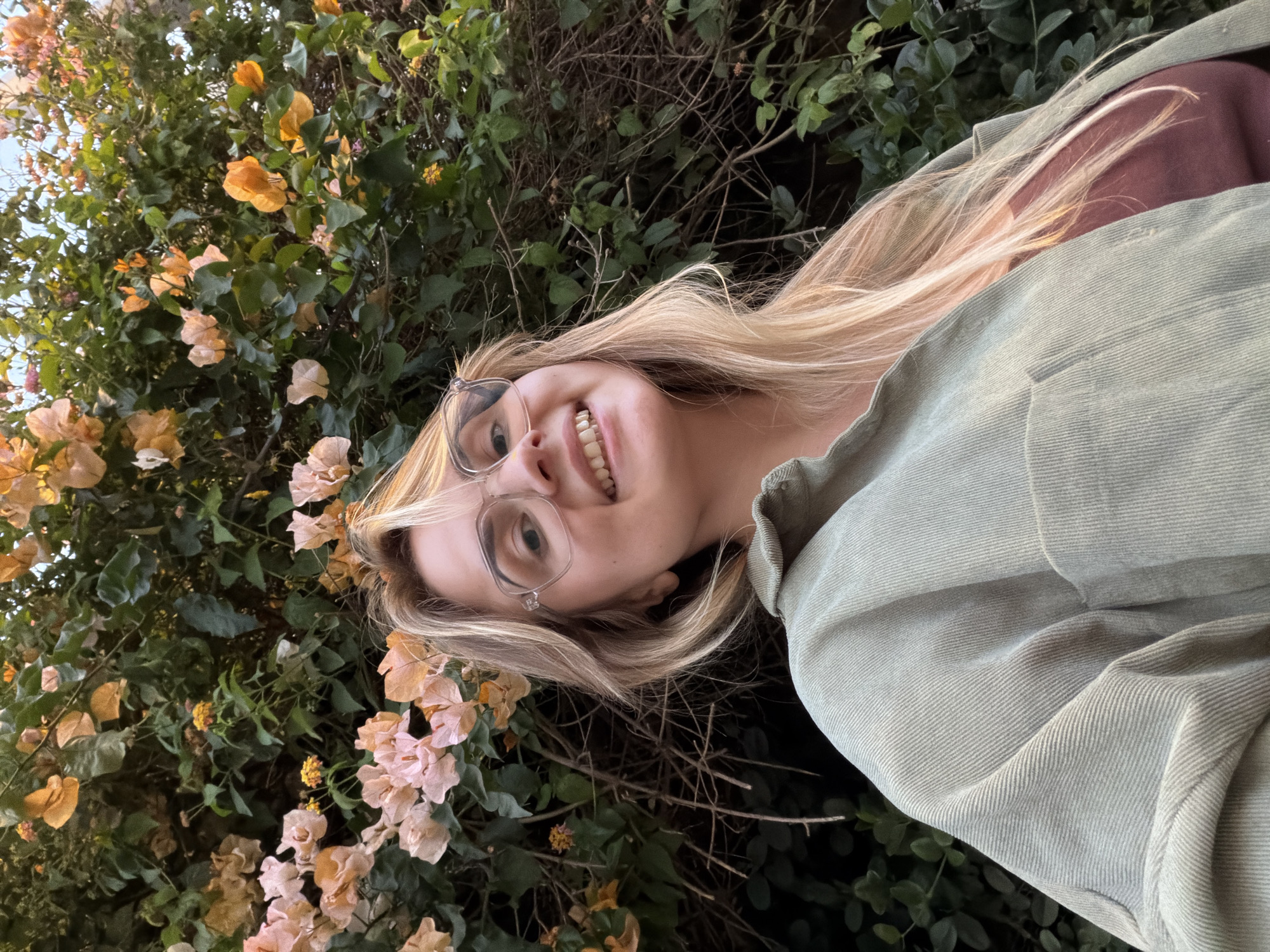 A young woman with long, blonde hair and clear-framed glasses smiles broadly at the camera in this outdoor, close-up shot. She is wearing a light sage-green, corduroy-textured jacket or shirt over a reddish-brown top. Her face is slightly angled, and she looks directly at the viewer with an open, happy expression, revealing her teeth.
The background is a dense tapestry of green foliage, likely a bush or climbing plant, heavily adorned with numerous small flowers. These flowers are primarily in shades of faded orange, light peach, and pale pink, resembling bougainvillea, which bloom abundantly in Mediterranean climates like Tel-Aviv, Israel. The soft, diffused light suggests either late afternoon or early evening, creating a warm and inviting atmosphere. No text is visible in the image. The overall mood is cheerful and natural.