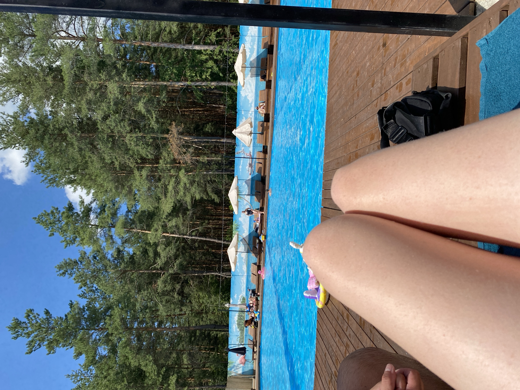 The image shows a view of a swimming pool area in Voronezh, Russia. In the foreground, a person's legs are visible, resting on a wooden deck. A black bag and a blue towel are also on the deck. The swimming pool is bright blue and appears to be outdoors. Several lounge chairs are set up around the pool, some with umbrellas providing shade. In the background, a dense forest of tall pine trees borders the pool area. A mural depicting a beach scene with a flamingo is visible on a wall behind the lounge chairs. Several people can be seen relaxing by the pool, some in lounge chairs and others near the water. A few inflatable toys are floating in the pool. The sky is a clear blue with a few white clouds.