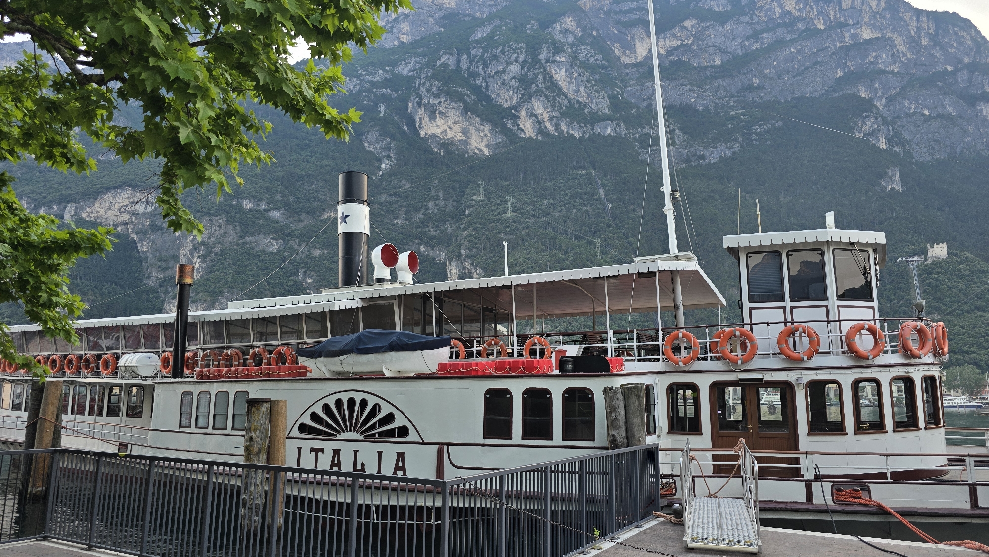 A paddle steamer named "ITALIA" is docked at a waterfront in Riva del Garda, Italy, with a steep, tree-covered mountain range in the background. The ship is white with a dark red trim and features multiple life preservers, a large smokestack with a star emblem, and a covered upper deck. Green foliage from an overhanging tree frames the left side of the image. The water is dark and calm, and a metal railing runs along the edge of the dock. The scene is bathed in soft, diffused light, suggesting it might be overcast or early morning/late evening. No people are visible in the frame.