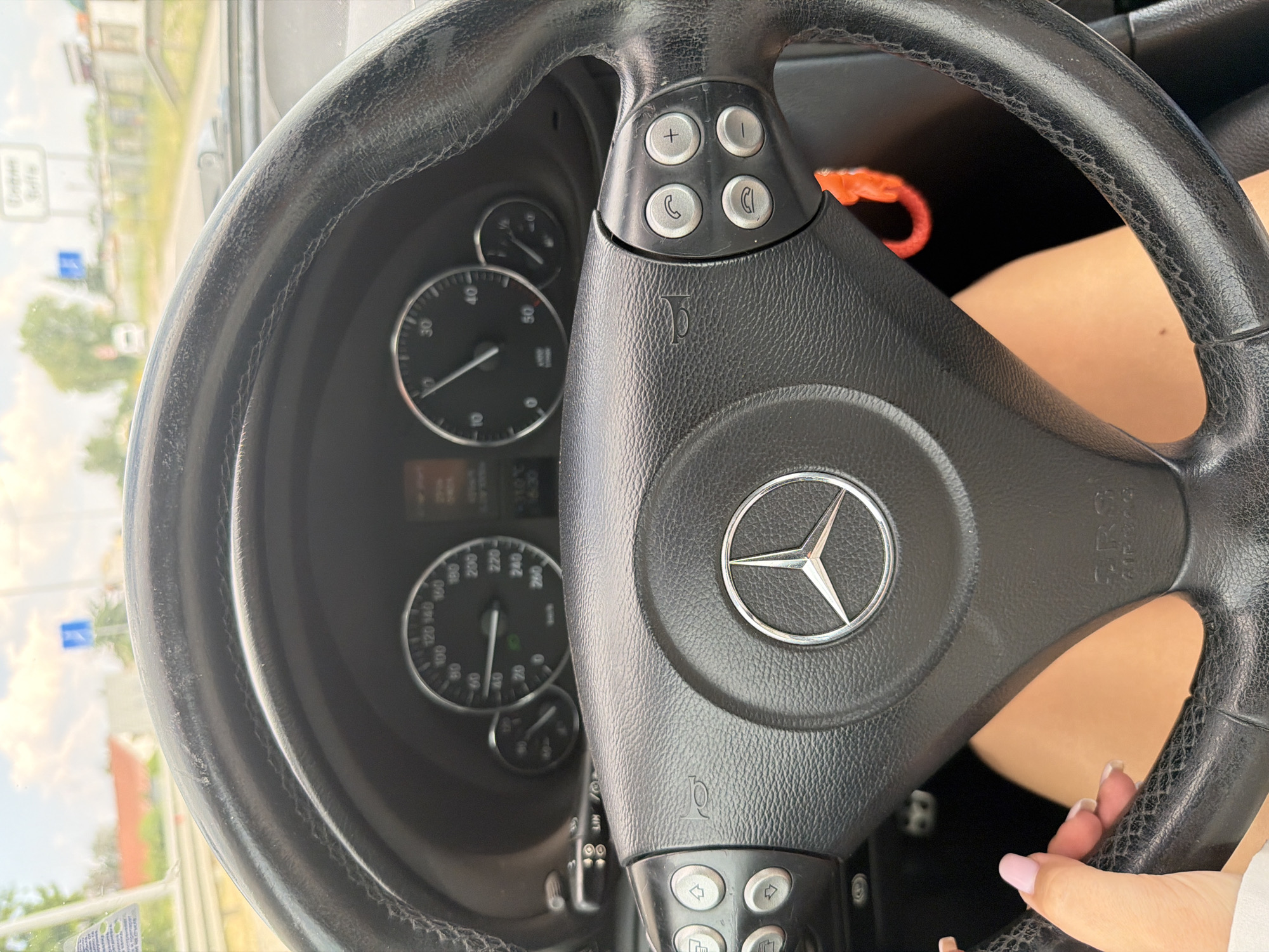 The image displays a first-person, eye-level view from the driver's seat of a Mercedes-Benz vehicle. The central foreground is dominated by a black, leather-wrapped steering wheel, prominently featuring a chrome Mercedes-Benz star emblem. The steering wheel's leather exhibits significant wear, characterized by cracking, peeling, and visible stitching deterioration across its upper and side rims. Multi-function control buttons are present on both the left (menu navigation) and right (telephone and volume control) spokes. An orange fabric lanyard or charm is visible hanging below and to the right of the steering column.
The driver's left hand, adorned with light pink polished fingernails, is positioned gripping the steering wheel at the nine o'clock position. A bare leg is visible between the lower spokes of the steering wheel.
Through the upper part of the steering wheel, a portion of the instrument cluster is visible. The speedometer indicates a vehicle speed of approximately 40 km/h, while the tachometer registers an engine speed of roughly 1500 RPM. A digital display is also partially visible within the cluster.
The external environment, viewed through the windshield, depicts a daytime scene under a cloudy sky. A paved road extends into the distance. To the left of the road, a metal guardrail and green vegetation are visible, along with a blue square road sign. To the right, blurred structures, including buildings and trees, line the roadside. A distant, highly blurred road sign with illegible text is also present. This scene is located in the city of Mramor, Bulgaria.