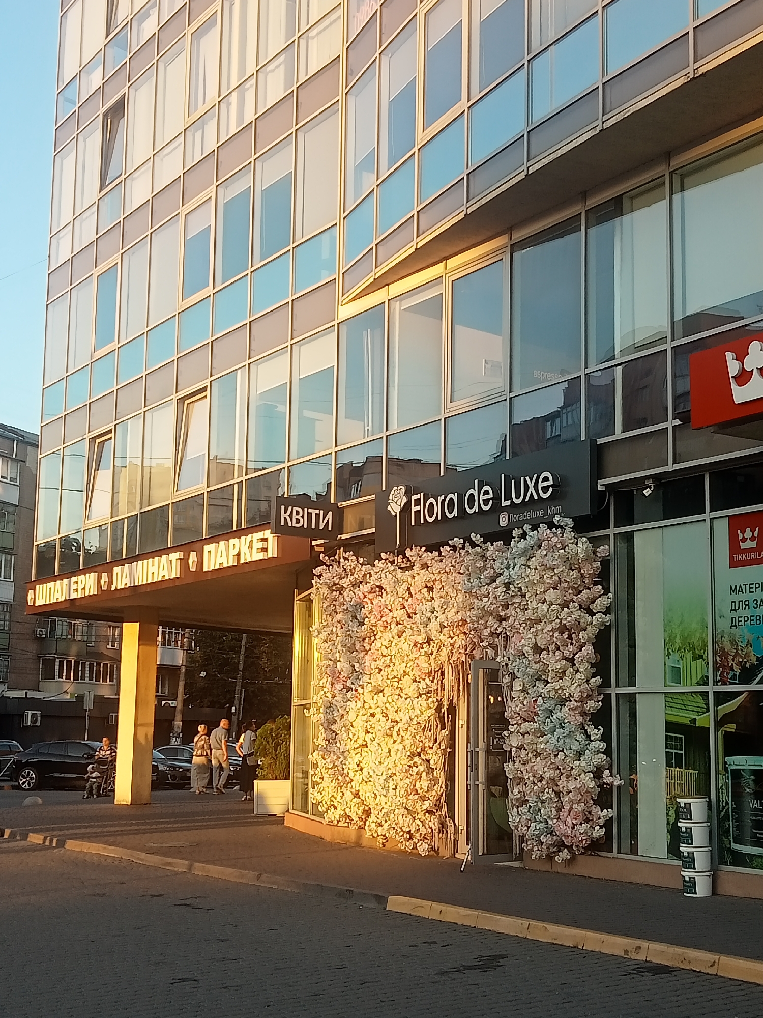 The image shows the exterior of a commercial building with large glass windows and a sign that reads "Flora de Luxe". The building appears to be in a city, as evidenced by the street and parked cars in the foreground. A group of people are walking on the sidewalk in front of the building, and a person is pushing a stroller. There are also several large, decorative floral arrangements outside the entrance to the "Flora de Luxe" establishment. The sun is setting, casting a warm glow on the scene.