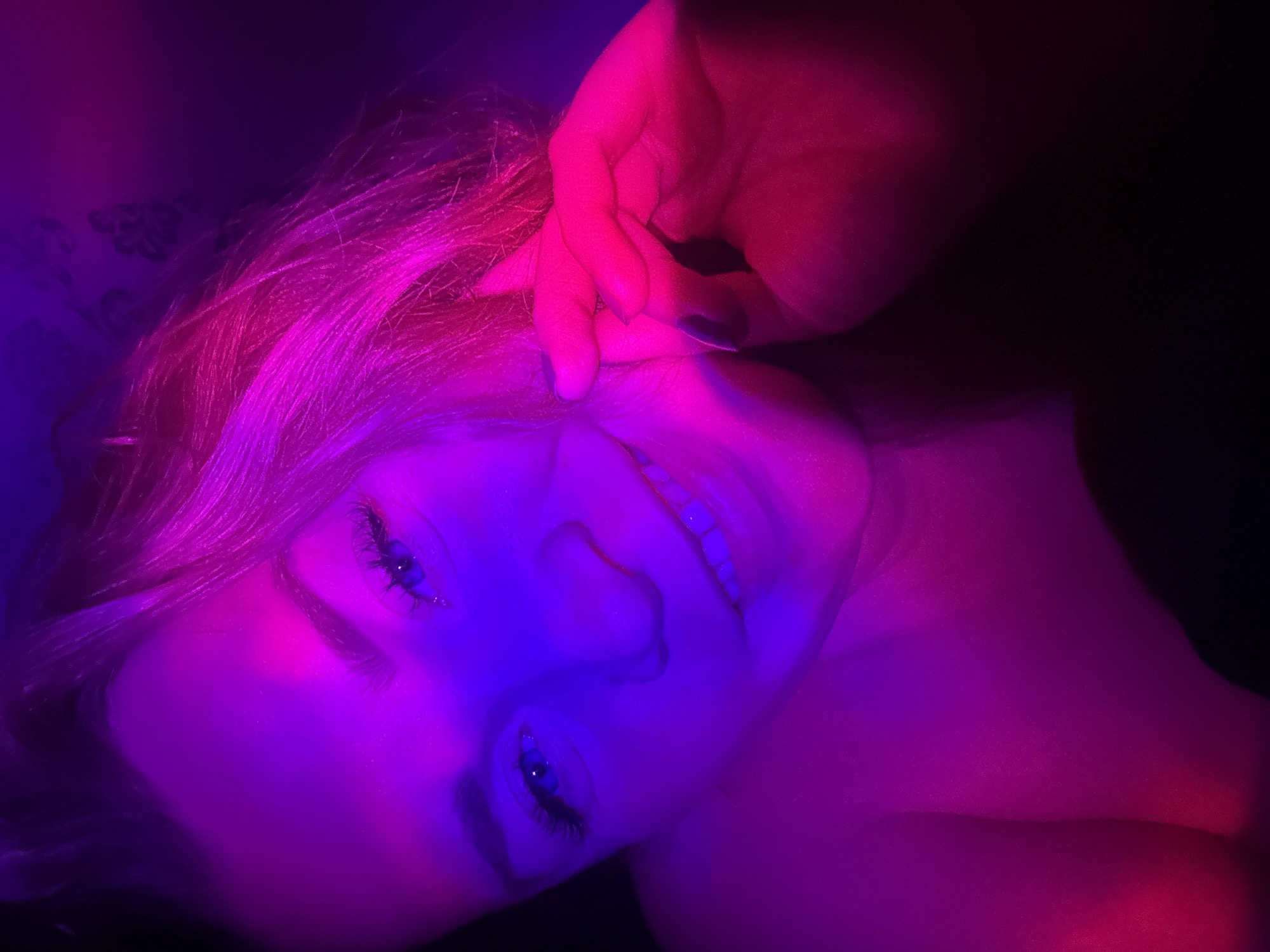 The image presents a close-up, low-angle portrait of a light-haired individual, likely female, illuminated by distinct dual-color lighting. The subject's face is angled slightly upward and to the right, occupying the central frame. The left side of the face, including the eye, and the upper left portion of the torso, are bathed in vivid blue and purple light. The right side of the face, hair, and a visible hand are illuminated with intense pink and magenta light.
The individual has blue eyes with prominent, dark eyelashes and a slight, engaging smile, revealing upper teeth. Light-colored hair, appearing vibrant pink-magenta due to the lighting, frames the right side of the face and extends towards the upper right corner. A left hand, featuring dark nail polish on several nails, is gently placed near the subject's right ear or temple, with fingers slightly splayed. The visible torso appears bare-shouldered. The background is dark, out of focus, and indistinct, showing subtle, unidentifiable patterns in the upper right, consistent with an indoor setting. No specific geographical or contextual identifiers are present within the image to determine the location, including the city of Veliky Novgorod, Russia.