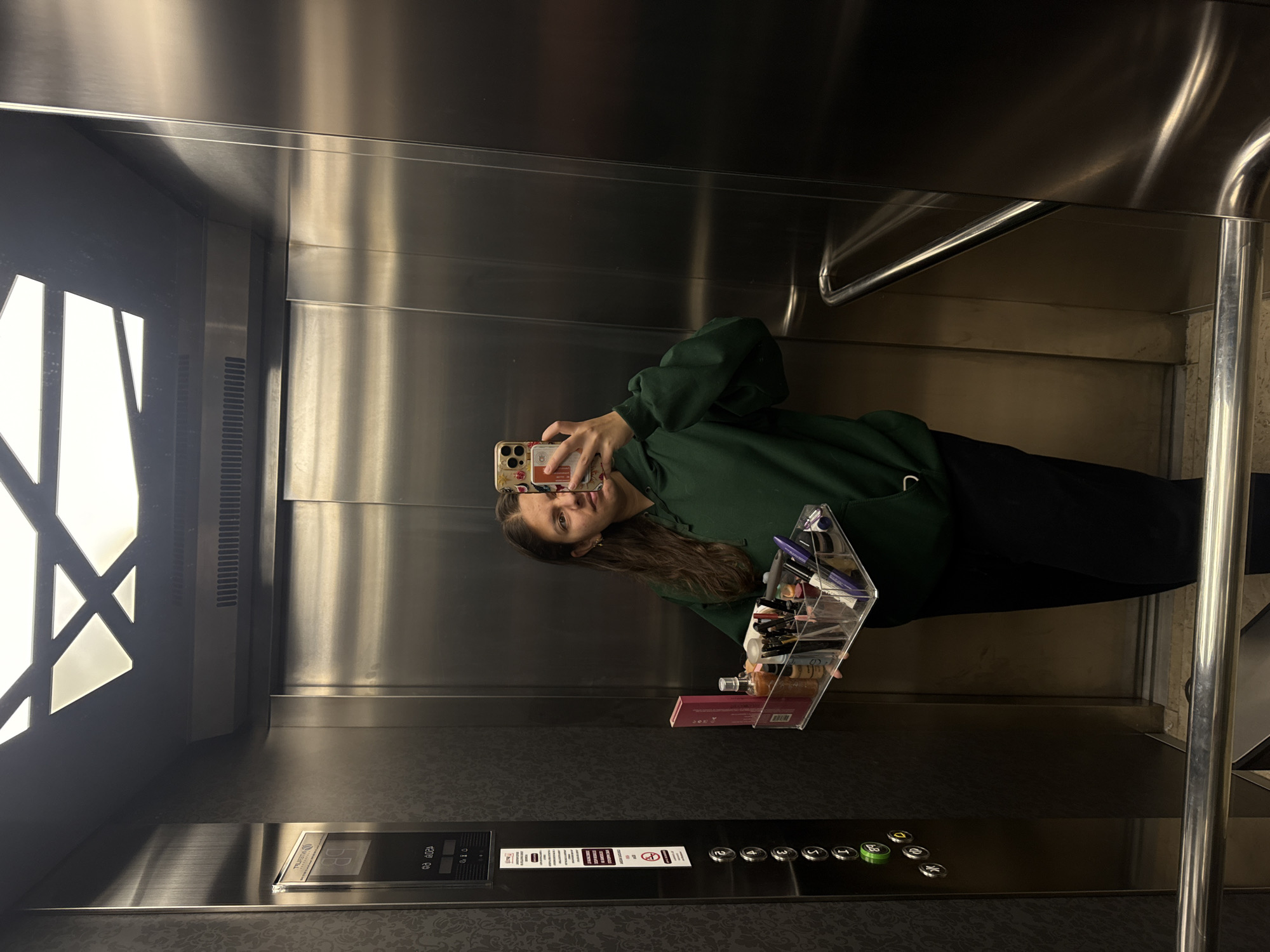 A young woman is taking a selfie in an elevator. She is wearing a green hooded sweatshirt and black pants. She is holding her phone in her right hand and a clear acrylic makeup organizer filled with makeup products and a pink box in her left hand. The elevator has stainless steel walls, a patterned floor, and a panel with buttons. The buttons are black with green accents. The elevator also has a decorative light fixture on the upper left side. There is a stainless steel handrail on the right side of the elevator. The location is unknown as the image does not provide information about San Bernardino Tlaxcalancingo, Mexico.