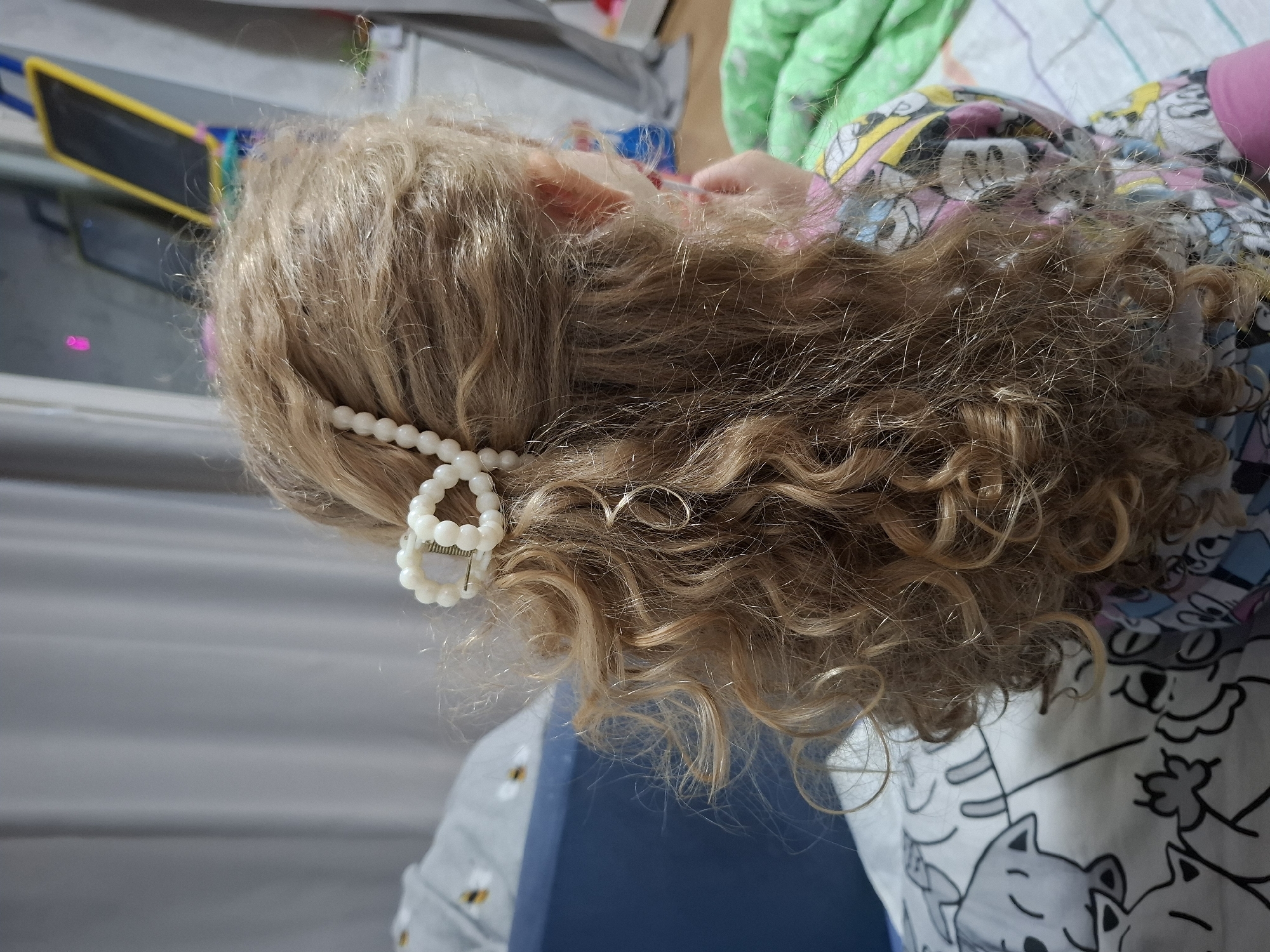 A close-up, eye-level shot shows the head and shoulder area of a child with curly, blonde hair. The hair is adorned with a pearl-beaded headband and a white, circular hair clip. The child is wearing a colorful pajama top patterned with cartoon characters. The background includes a blurred blue and white object, likely a chair or furniture, and a section of a surface with a yellow and black object in the upper left corner. There's no discernible information to confirm the location is Chaikovsky, Russia, or any other specific location.
