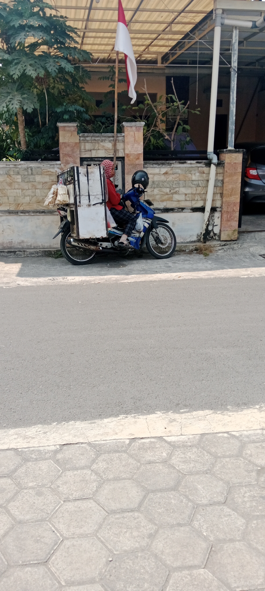 In Maguwoharjo, Indonesia, two individuals are seated on a blue and white motorcycle. The person in the front, wearing a red top and a patterned headscarf, is looking to the right. The person behind them is wearing a black helmet and a blue jacket, with a checkered pattern on their pants. The motorcycle is equipped with a large white cargo box attached to the rear, which appears to be carrying items wrapped in beige plastic. The motorcycle is parked on the side of an asphalt road, next to a sidewalk paved with hexagonal tiles. A stone wall with a brick accent runs behind the motorcycle. A flag, red on top and white on the bottom, is visible on a bamboo pole above the scene. The environment suggests a sunny day, with shadows cast on the road and sidewalk. In the background, a car is partially visible and some trees and a building can be seen.
