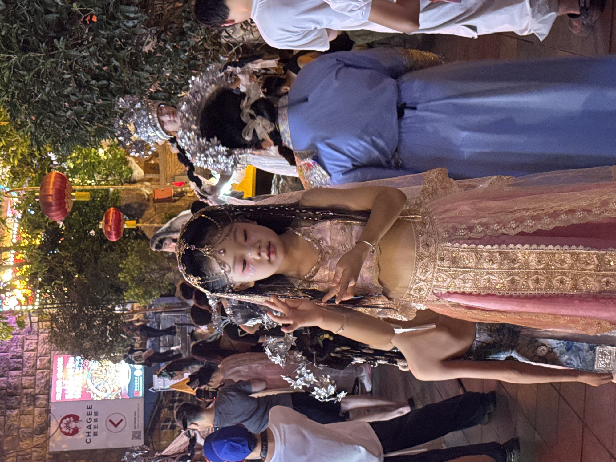 In Yuzhong District, China, at night, a young girl with face paint and elaborate headdress stands in the foreground, adorned in a shimmering pink and gold outfit with exposed midriff and intricate jewelry. She is performing a hand gesture. Behind her, a person in a blue garment with a braided bun stands with their back to the camera, also wearing a decorative headdress. To the left, a young person in a blue hat and white t-shirt is visible. The background depicts a street scene with illuminated trees, red lanterns, and a brick wall, suggesting a festive event or market. Other people in casual attire are scattered throughout the scene.