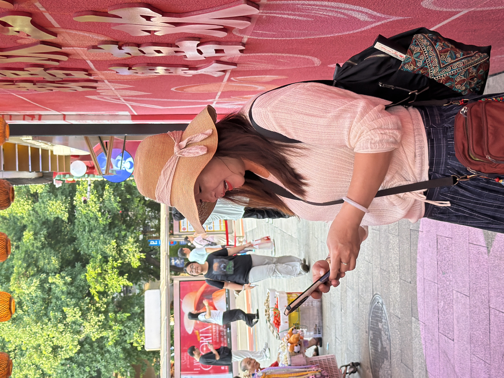 A woman wearing a straw hat and a light pink sweater stands on a city street in Yuzhong District, China, looking at her smartphone. She has long brown hair and is wearing a blue striped skirt and a white bracelet. A black backpack is slung over her shoulder, with a decorative pouch peeking out. Behind her, other people are visible walking and browsing at market stalls. The background features a bright pink wall with ornate gold lettering, lush green trees, and orange hanging lanterns. The street is paved with grey and pink tiles.