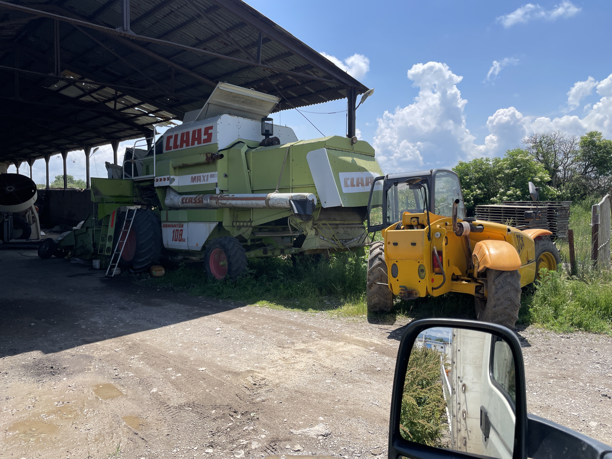 An outdoor agricultural scene in Gorni Bogrov, Bulgaria, is depicted on a bright, sunny day with a clear blue sky adorned with large, fluffy white cumulus clouds.
In the foreground, a large green and white Claas combine harvester is partially parked under an open-sided shed with a corrugated metal roof, casting a shadow over the foreground ground. Visible text on the combine includes "CLAAS," "MAXI," and "DOMINATOR 108 SL." A red ladder leans against the rear wheel of the combine. To the right of the combine, a yellow JCB telehandler is parked on the unpaved ground, which is a mix of dirt and gravel. Several muddy puddles are present on the ground, indicating recent wet conditions.
Beyond the vehicles, green grass and weeds grow around the machinery and at the edges of the shed. Further in the background, a line of lush green trees and shrubs is visible under the bright sky. The shed itself has a visible metal framework supporting the roof, with glimpses of the sky through gaps in the structure.
The image is taken from the perspective of someone inside a vehicle, with a side-view mirror occupying the bottom right corner of the frame. The mirror reflects a portion of a white truck or van and some green foliage. No people are visible in the scene, and the heavy machinery appears to be stationary, possibly stored or awaiting maintenance.