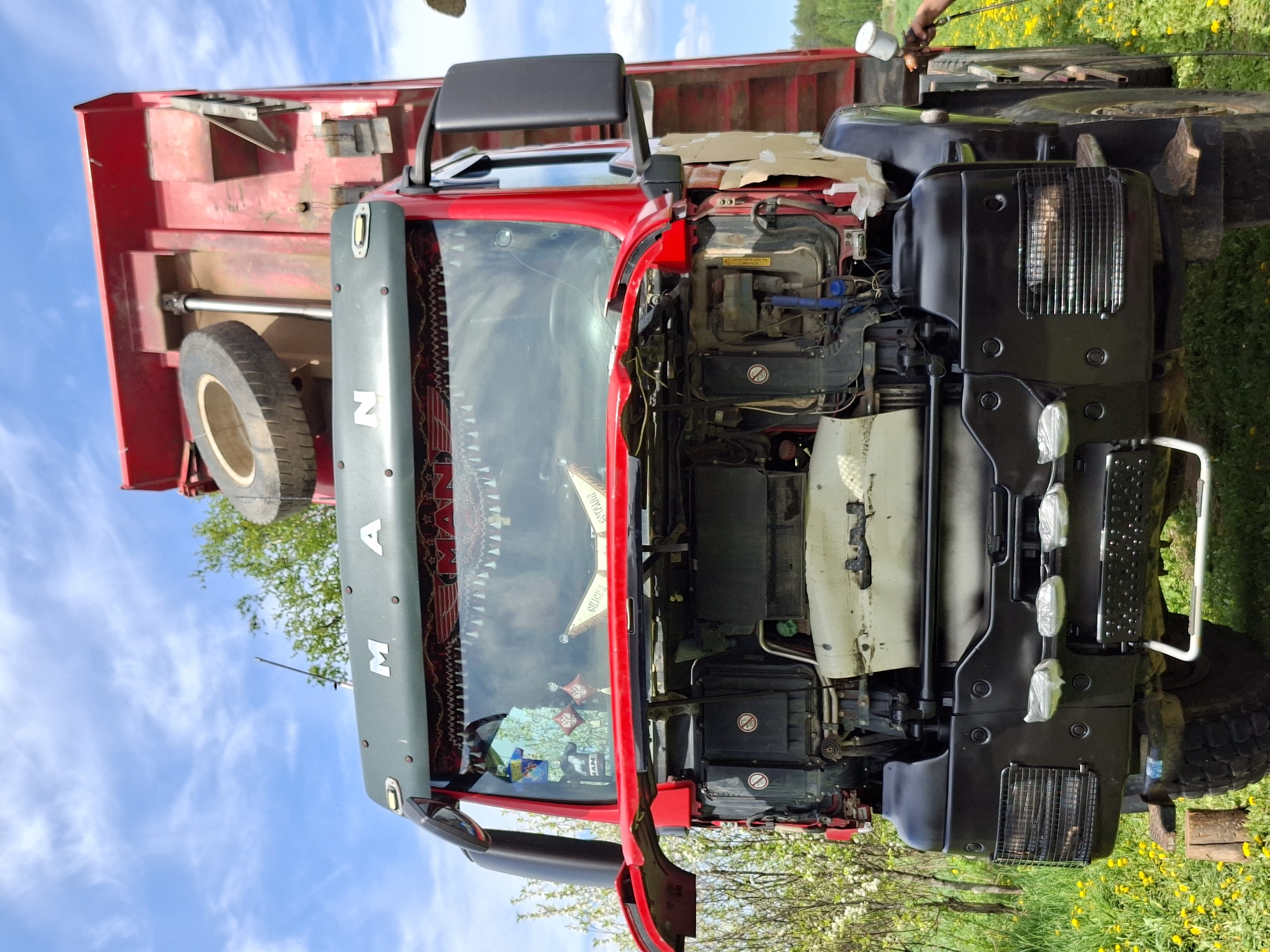 The image depicts a red MAN truck in an outdoor setting, likely a field, with dandelions visible in the foreground. The truck's hood is open, revealing the engine components. A spare tire is mounted on the back of the truck bed. A person is seen painting the front of the truck black. The sky is visible in the background, showing a partly cloudy day. The location is not identifiable as being in any specific city, Unknown, Unknown.
