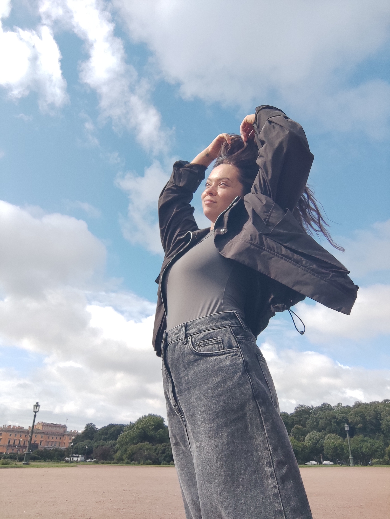 A woman is captured from a low angle, looking upwards and to the right, with her hands in her dark hair against a bright blue sky with scattered white clouds. She is wearing a dark grey, high-waisted denim skirt and a dark grey long-sleeved top, with a black utility jacket partially open over the top. The woman's stance and pose suggest a moment of freedom or contemplation. In the background, a wide, light-colored path leads towards a line of green trees. To the left, a large, ornate building with a light orange facade and multiple windows is visible, and a traditional lamppost stands in front of it. This scene appears to be set in a park or an open area within the city of Saint Petersburg, Russia, likely in the vicinity of a palace or grand estate given the architecture.