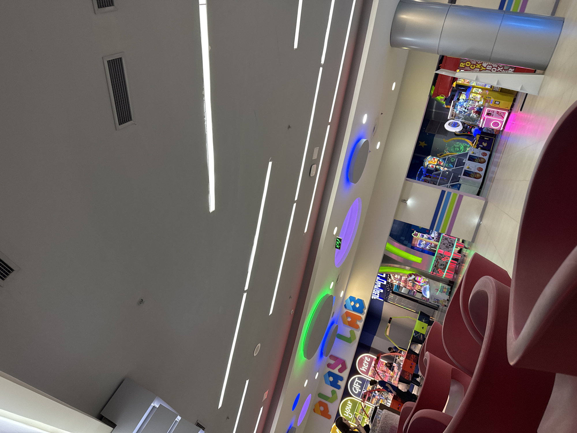 This image depicts a vibrant indoor entertainment area, likely an arcade or family play zone within a shopping mall. The perspective looks upwards and across the brightly lit space.
In the foreground, several red, curved, contemporary seating units are visible, partially obscuring the view of the background. A prominent white wall features colorful circular cutouts with LED lighting in shades of blue, green, and purple. Large, illuminated letters spelling "PLAY LAB" are fixed to this wall, identifying the area. A small green "EXIT" sign is also visible in the distance above the "PLAY LAB" sign.
The background reveals numerous arcade games, including claw machines (one clearly labeled "GIFT here" and "your"), basketball shooting games, and a large red and white machine that appears to be a "ROCKY BOXER" or "ROCK BOX" game. Other brightly lit game machines line the walls. A few people are visible in the arcade area, seemingly engaging with the games or observing. A tall, cylindrical, metallic-clad pillar stands on the far right.
The ceiling is white and features multiple long, linear fluorescent light fixtures, providing ample illumination. Several rectangular air vents are also integrated into the ceiling. The overall impression is a modern, clean, and lively public space, consistent with entertainment centers found in locations such as Yerevan, Armenia.
