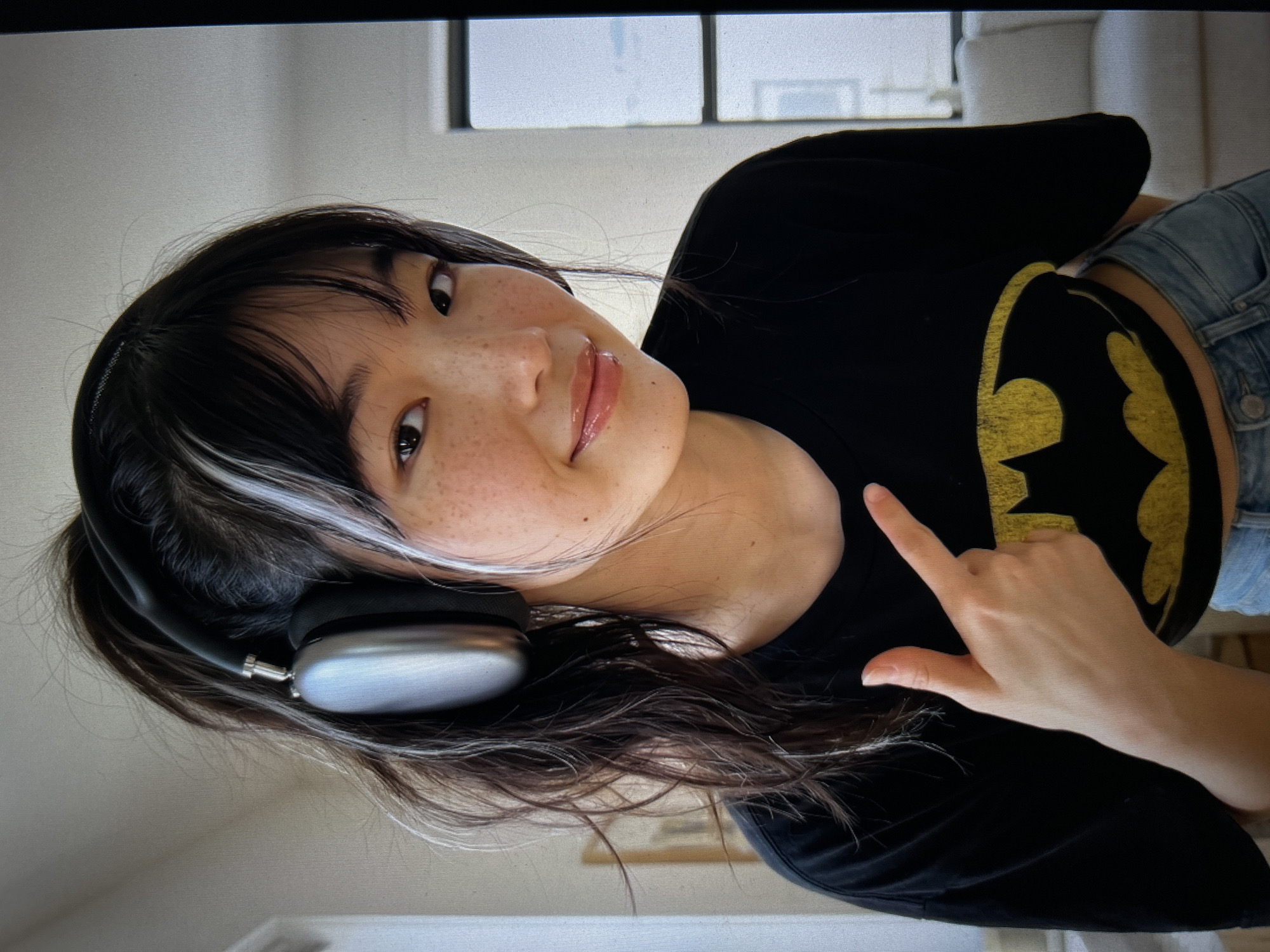 A young Asian woman with freckles and bangs, highlighted with streaks of white in her dark hair, is looking at the camera and smiling. She is wearing a black t-shirt with a yellow Batman logo and light blue jeans. She has on a pair of silver headphones. Her right index finger is pointing towards the camera. The background is out of focus but appears to be an indoor setting with a couch and window. The scene is well-lit.