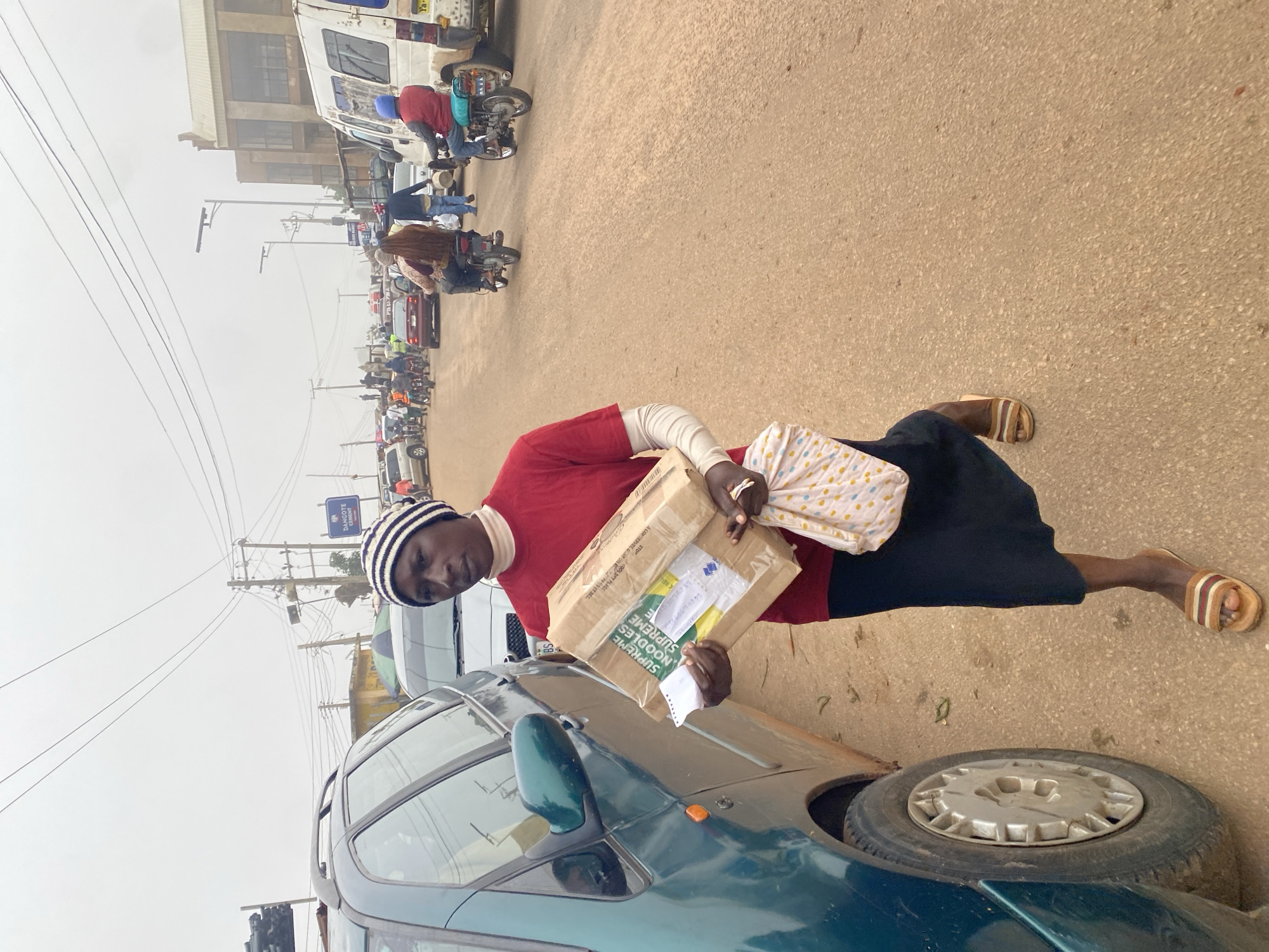 A Black woman walks on a dusty road, carrying a cardboard box and a patterned cloth bag. She wears a red long-sleeved shirt, a black skirt, a black and white striped beanie, and striped sandals. The box, labeled "SUPREME NOODLES SUPREME," is held in her right arm, while the cloth bag is clutched in her left hand. To her left, a green car is parked. In the background, a busy street with traffic, motorcycles, and buildings suggests an urban environment. The sky is overcast, casting a diffused light on the scene, indicative of a cloudy day. This is an outdoor scene in Gura Top, Nigeria.