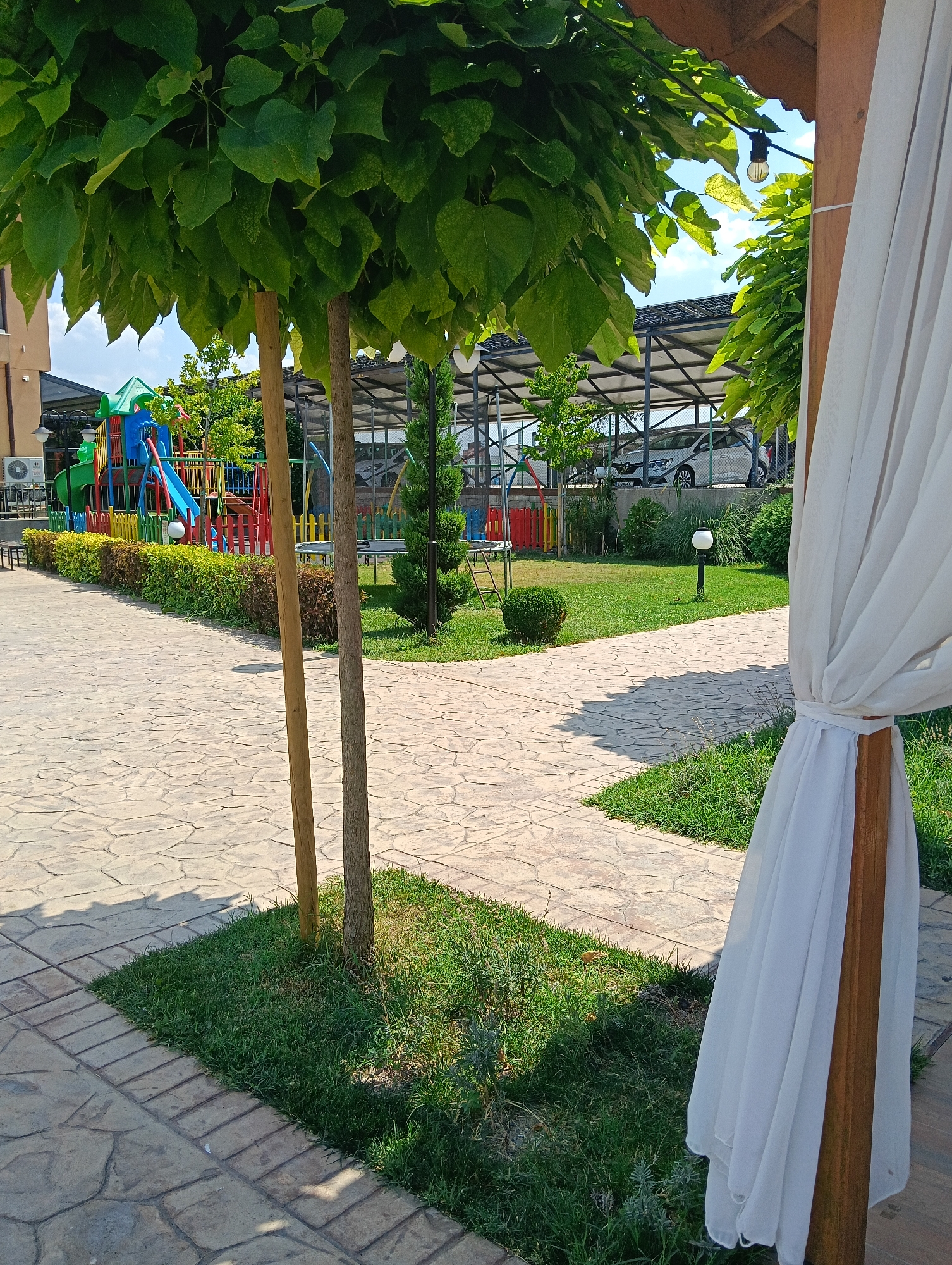The image shows a view of a playground area with a paved walkway and a covered seating area with white drapes. In the background, a white car is parked under a covered structure. The playground equipment is colorful, with a slide, swings, and climbing structures. There are trees and green grass surrounding the playground. The scene is well-lit, suggesting it is daytime. The location is specified as the city of Radievo, Bulgaria.