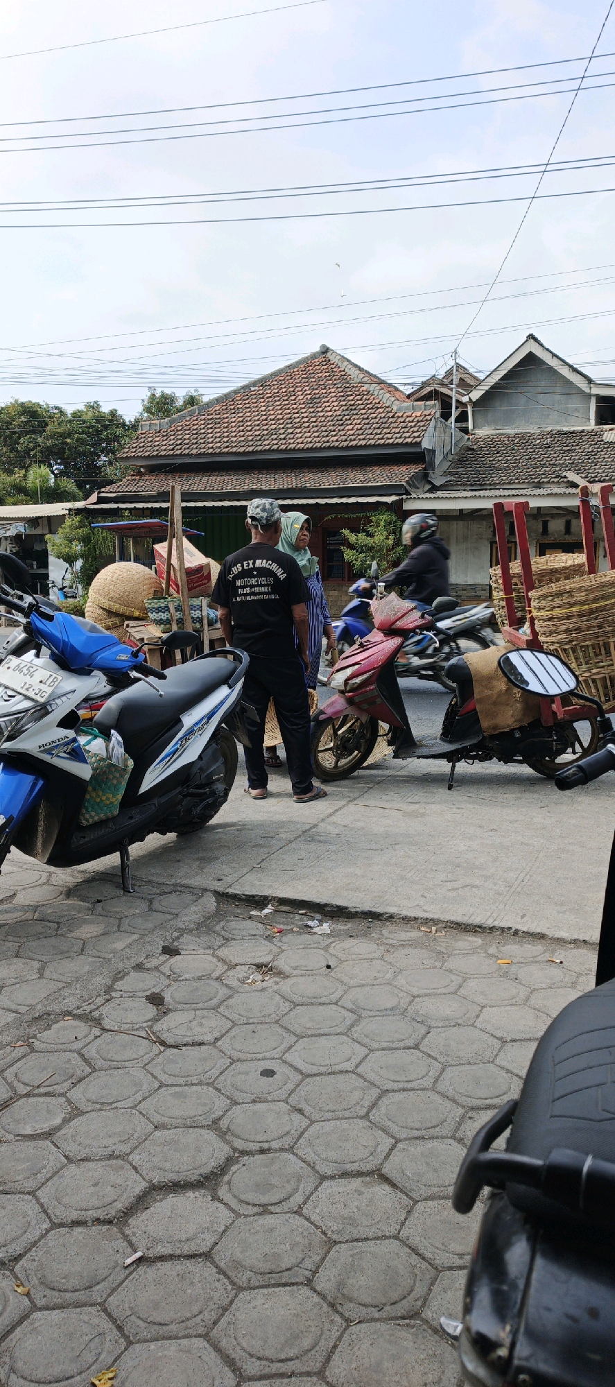 In Kasiyan, Indonesia, on a paved road with hexagonal concrete tiles, three individuals are visible. A man wearing a black t-shirt with "PLUS EX MACHINA MOTORCYCLES" printed on the back and a camouflage baseball cap stands facing away from the camera, engaged in conversation with a woman wearing a light green hijab and a blue and white striped dress. To their right, another person, likely a man, wearing a helmet and a black jacket, is riding a red scooter that is in motion, blurring the image. Several parked motorcycles are present, including a white and blue Honda scooter in the foreground. Behind the people, there are houses with red tiled roofs and what appears to be a market stall with woven baskets and merchandise. Utility wires crisscross the sky above.