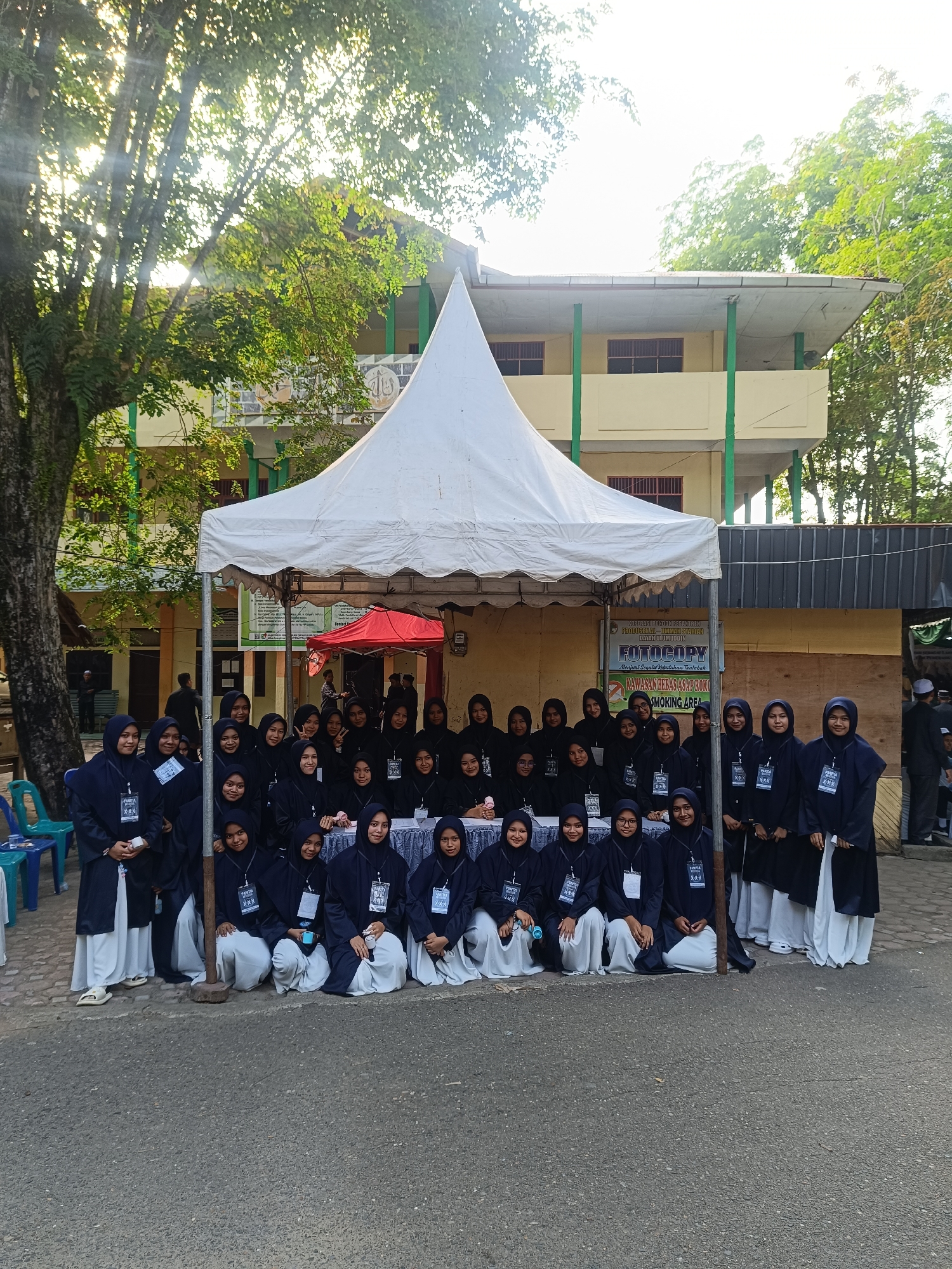 The image captures a group of approximately 25 young women, all dressed in matching uniforms: dark blue jackets and hijabs over white skirts, each wearing a badge with the text "PANITIA XXX." They are posed in two rows, seated and standing, in front of a white tent bearing a sign, in Bahasa Indonesian, stating "Koperasi Pondok Pesantren Produsenal-Ummah Syariah Dayah Ulumuddin, Fotocopy, Menjual Segala Kebutuhan Thalabah, Kawasan Bebas Asap Rokok / Smoking Area." A building of two stories with a yellow facade and green pillars serves as a backdrop. The image is taken outside, possibly in the city of Lhokseumawe, Indonesia, given the language on the sign and the attire of the subjects. A paved road is visible in the foreground, and a large tree is on the left side of the frame.