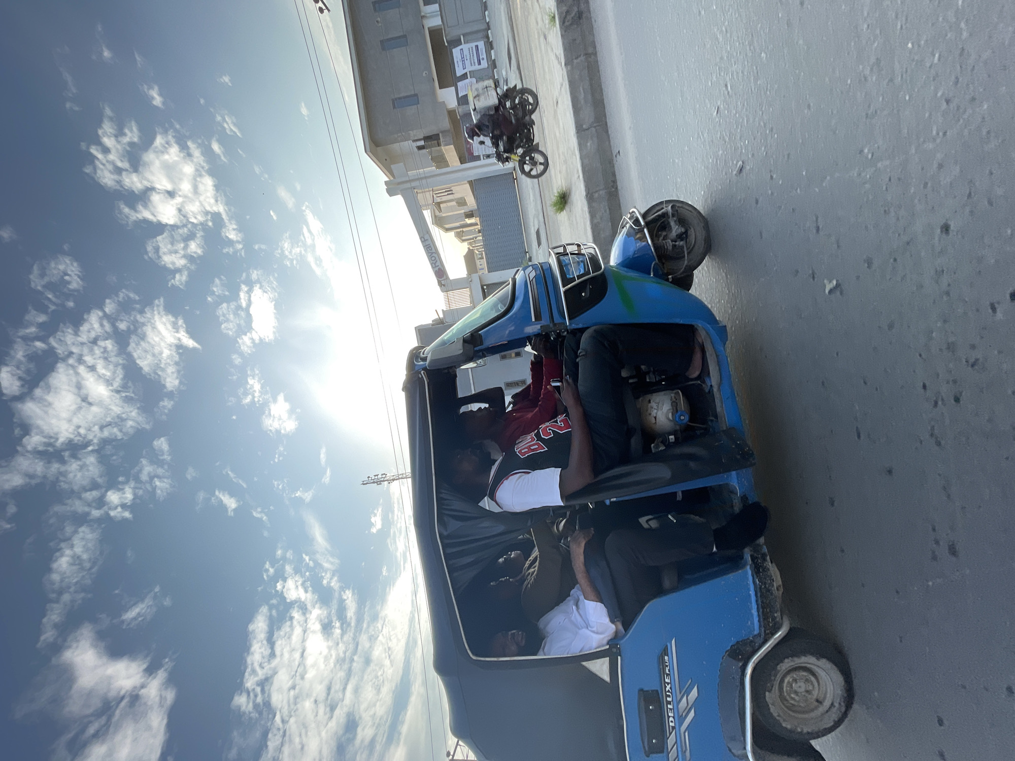 The image shows a blue auto-rickshaw with several people inside, traveling on a paved road. The auto-rickshaw has a covered passenger area and a visible engine at the back. There are at least four people visible in the auto-rickshaw, with some appearing to be seated and others possibly standing or in transit. One person in the front is wearing a red basketball jersey with the number 23. The scene appears to be in a semi-urban or urban environment with modern buildings in the background. A motorcycle is visible in the background, with its front wheel raised, suggesting a stunt or playful activity.
The sky is bright with scattered clouds, and the sun is shining, indicating it is daytime, likely late morning or afternoon. The lighting suggests a clear day. The road surface appears to be asphalt, and there are some white markings visible, possibly lane dividers or road signs. The overall atmosphere seems busy and dynamic, with the auto-rickshaw in motion.
Visible text on the auto-rickshaw includes "DELUXE PLUS" and some unreadable lettering. There is also a sign on a building in the background with text that is difficult to decipher. The location is contextually identified as Ado, Nigeria. The people's expressions are not clearly discernible due to the image quality and angle, but the activity suggests a common mode of transportation.