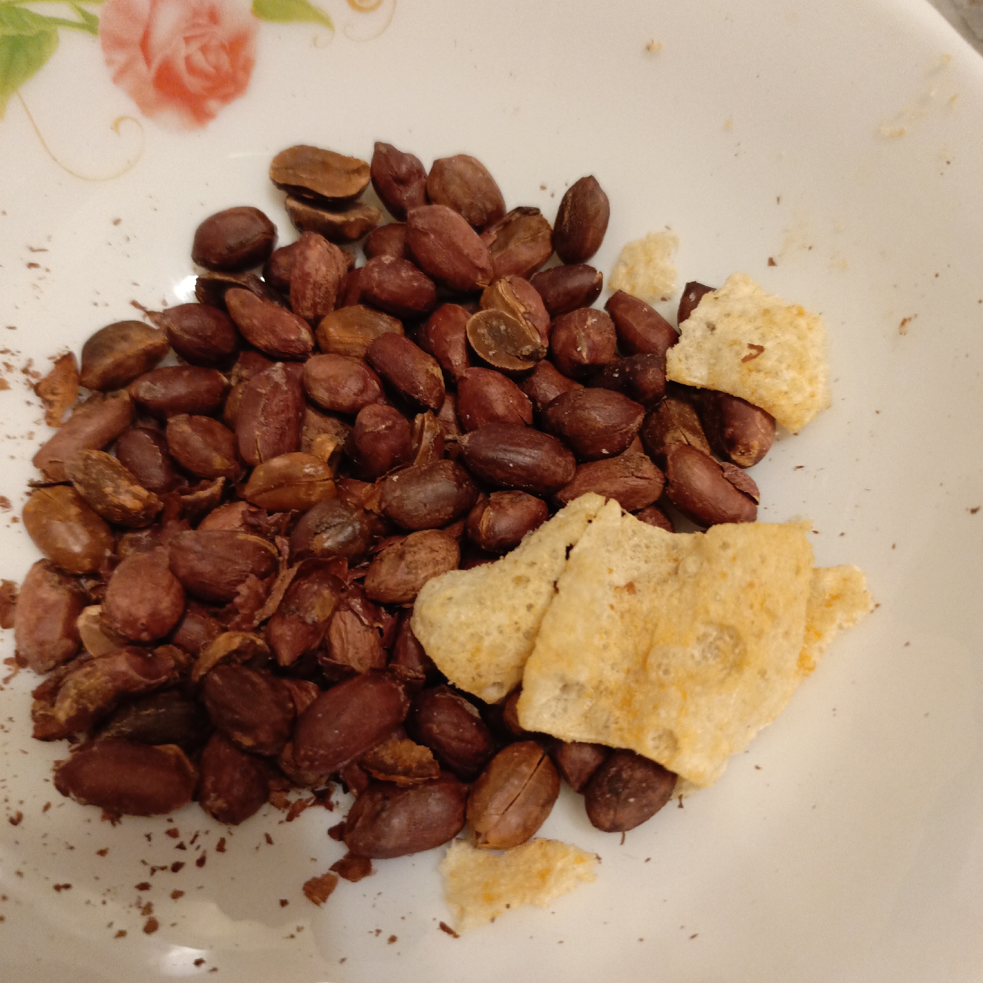 A white plate with a red flower design contains a pile of roasted peanuts and fragments of what appears to be a flat, rectangular crispy snack, possibly made from rice or tapioca flour. Small crumbs are scattered around the peanuts. The location is unknown.