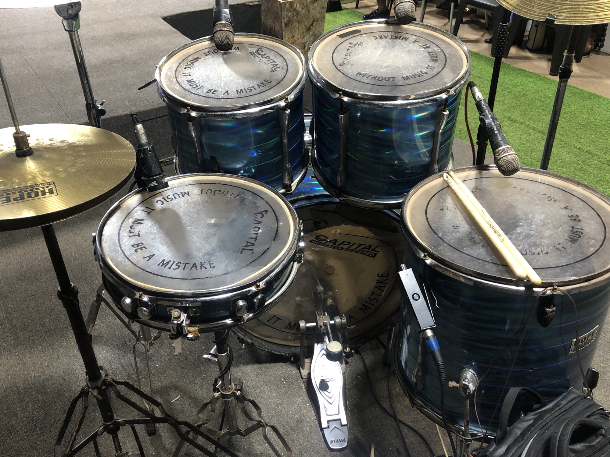 A drum kit, featuring four drums and a cymbal, is positioned on a floor with mixed surfaces, part of which appears to be carpeted. The drums are dark blue with silver rims, and each drumhead is inscribed with the words "CAPITAL WITHOUT MUSIC IT MUST BE A MISTAKE". Microphones are attached to several of the drums. A pair of drumsticks rests on the rightmost drum. The drum kit includes a Tama bass drum pedal. A "Hope" cymbal is mounted on a stand to the left. The context and location within the city of Onitsha, Nigeria, are not evident from the provided image.