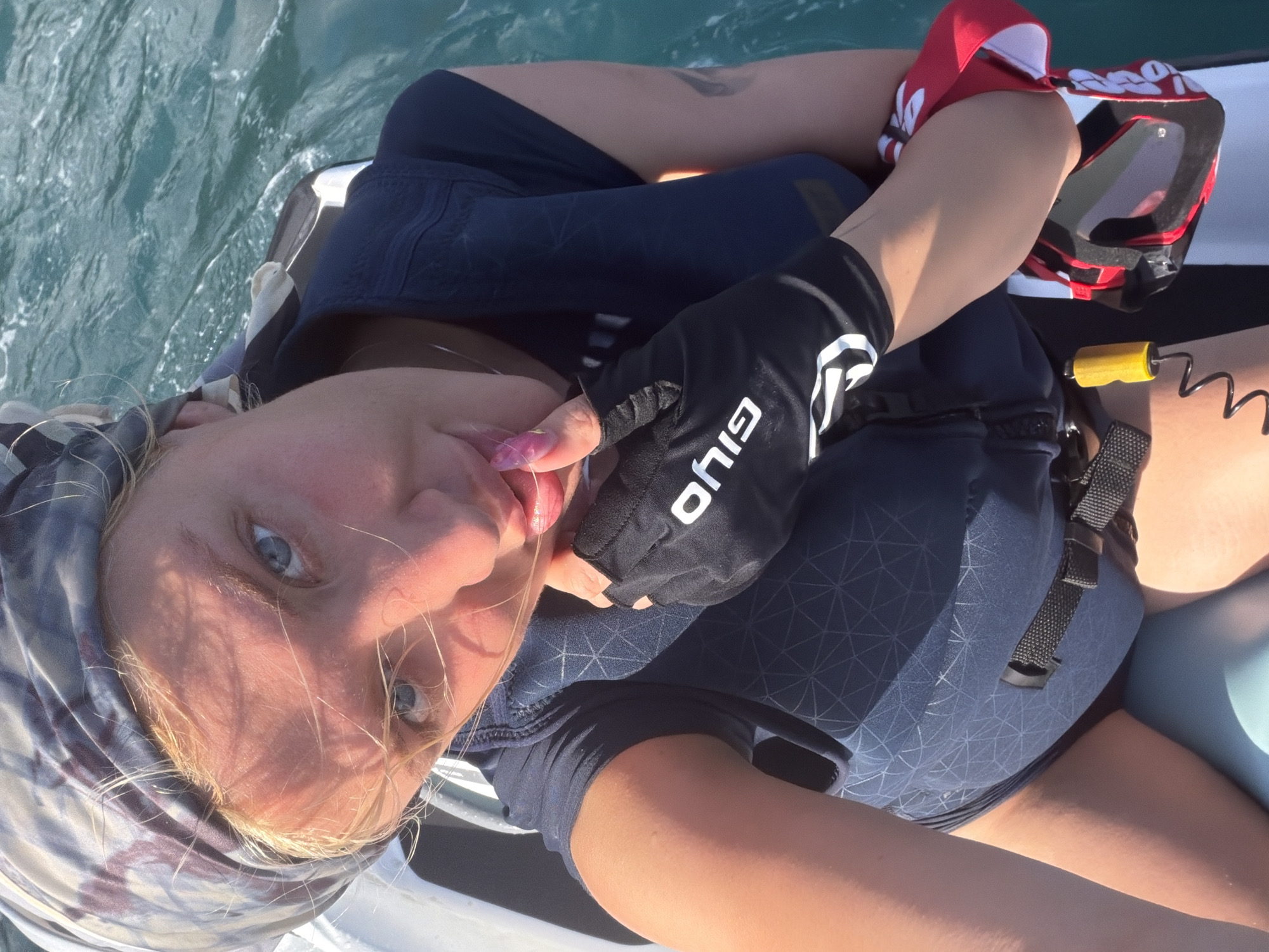 A woman with blonde hair and blue eyes, wearing a bandana, a navy blue life vest, and black gloves, is taking a selfie while riding a jet ski in the ocean. The water is a vibrant turquoise and appears choppy, suggesting movement. Her right hand, adorned with pink nail polish, is touching her face. A pair of goggles with red and black straps is held in her left arm. The scene is outdoors, with bright sunlight casting shadows on her face and chest, indicating it is daytime. Based on the attire and activity, the location is likely a water sports area in Nusa Penida, Indonesia.
