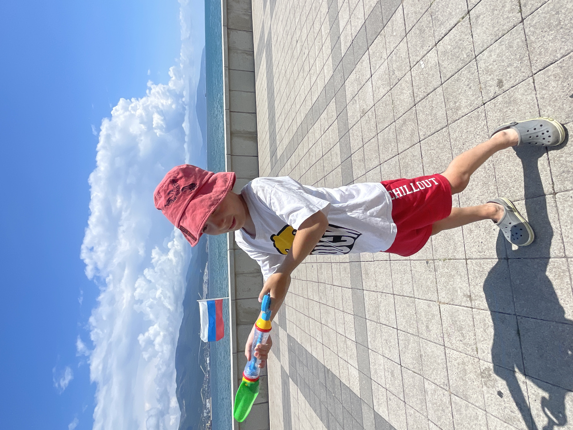 A young boy wearing a red hat, a white t-shirt with a yellow smiley face graphic, and red shorts, stands on a paved promenade near the sea. He is holding a plastic water gun, with a green nozzle and a clear body containing blue liquid. The promenade is tiled with light gray squares. In the background, a Russian flag flies from a pole. The sea is calm and a mountain range is visible in the distance. The sky is blue with scattered white clouds. The scene appears to be taken on a sunny day. The location is likely the promenade in Novorossiysk, Russia, given the presence of the Russian flag and the coastal setting.