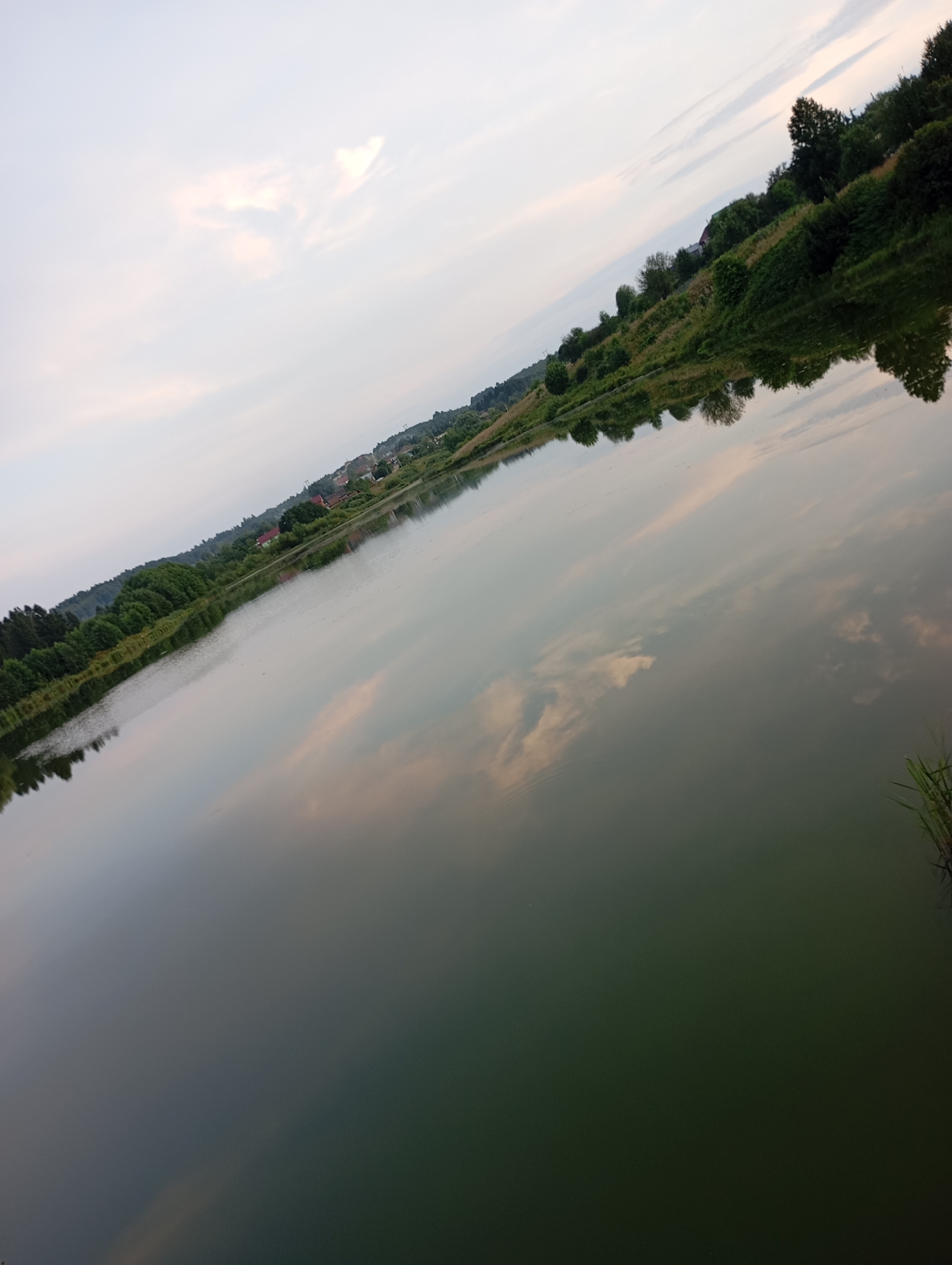 The image depicts a lake with a reflection of a tree-lined hill with some houses on its slope. The sky is overcast with some pale clouds. The water is dark and calm, reflecting the scene above. The perspective is from the water's edge, looking across the lake towards the hill. The image is taken at an angle, with the horizon tilted. There are no people or animals visible in the image. The scene is peaceful and natural, with a hint of human habitation on the hill. The location is identified as Фоки, Russia.