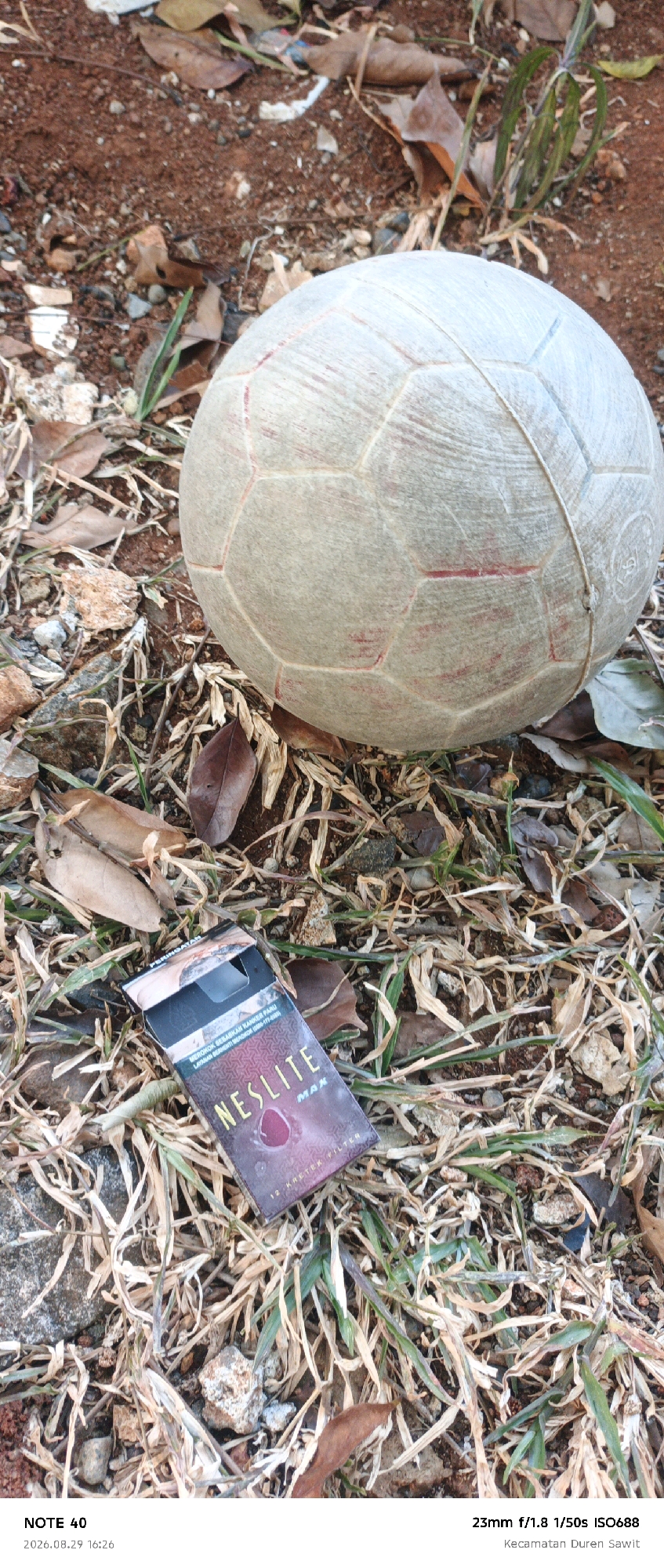 The image depicts an outdoor scene on the ground, likely in a park or a residential area. In the upper portion of the frame, a worn, off-white soccer ball with red stitching is visible. The ball rests on a surface of reddish-brown soil mixed with dried leaves, grass, and small stones. In the lower portion of the image, an open pack of "NESLITE MAX" cigarettes is placed on the ground, surrounded by dry grass and leaves. The cigarette pack has a dark purple design with the brand name in yellow lettering. The timestamp at the bottom of the image indicates the photo was taken on August 29, 2026, at 16:26. The location is specified as Kecamatan Duren Sawit, East Jakarta, Indonesia.