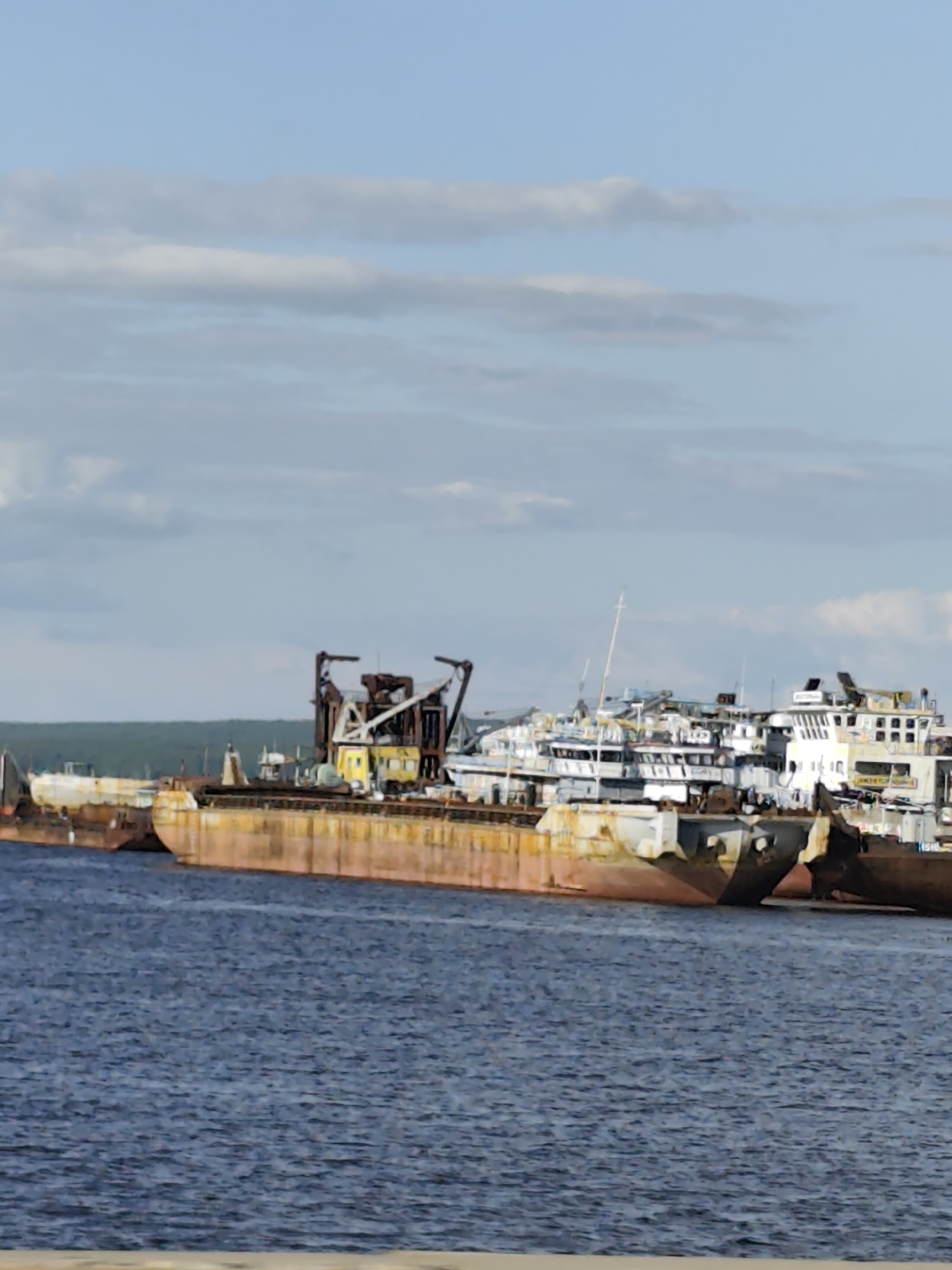 The image depicts a fleet of rusted, decommissioned barges and ships docked in the water, likely a ship graveyard or storage area. The vessels are aged, with visible signs of wear and tear, including corrosion and disrepair. In the background, a line of green trees borders the shoreline under a partly cloudy sky. The water is a deep blue, with a slightly choppy surface.
This scene is characteristic of the Chaikovsky Shipyard, a location in Chaikovsky, Russia, known for its ship repair and decommissioning activities on the Kama River. The yard has historically been a place where old vessels are brought for storage or dismantling.
