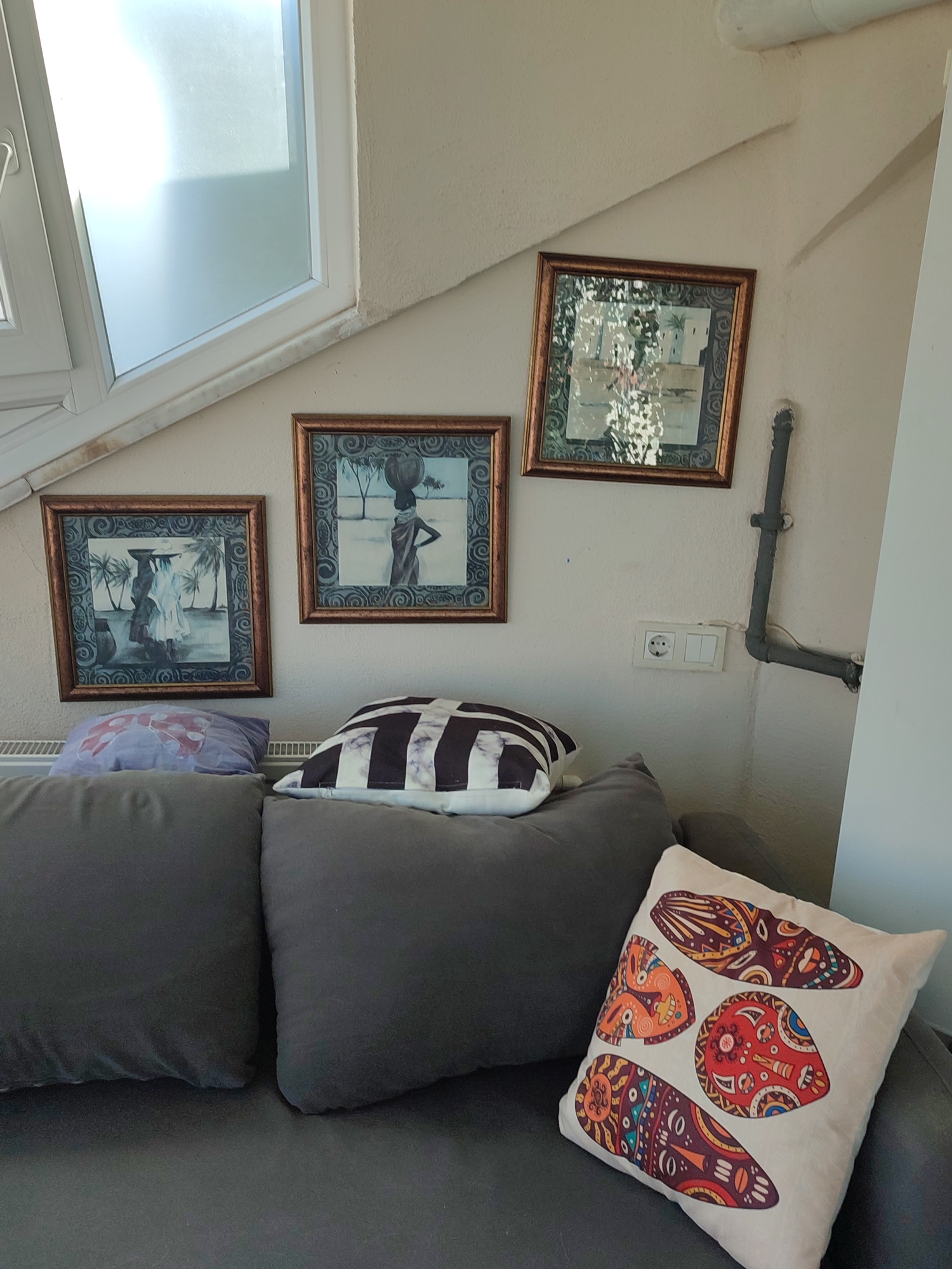 The image depicts an indoor scene featuring a gray sofa adorned with several pillows. Two framed artworks hang on a textured, off-white wall above the sofa. The artwork on the left shows two women carrying baskets on their heads in a tropical setting with palm trees. The artwork on the right is a framed print of a building with reflections of trees. Adjacent to the artworks is a wall-mounted electrical outlet with two switches. A dark gray pipe runs vertically along the wall next to the outlet. A radiator is visible below the pillows on the sofa. A large window with white framing occupies the upper left portion of the image. The overall impression is that of a living room. This image was taken in Üsküdar, Turkey.