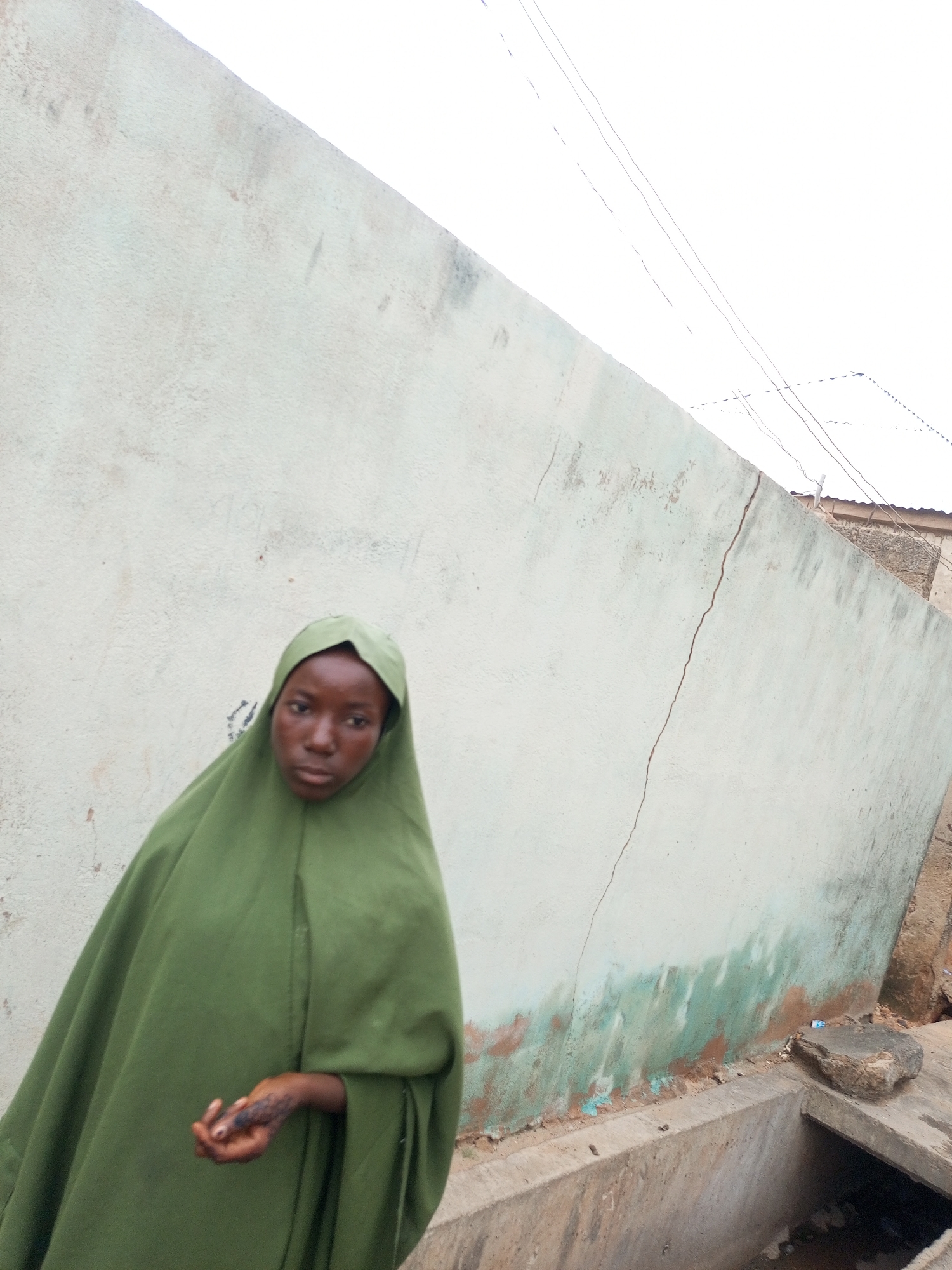 In an outdoor environment, a young person, wearing a green hijab that covers their head and shoulders, stands in front of a light-colored, cracked wall. The person's face is visible, with dark skin and hair. Their left hand is extended forward, showing a dark pattern on the palm, possibly henna. The wall behind them has patches of green and brown discoloration near the bottom. A concrete channel runs along the base of the wall, and a stone rests on its edge. Overhead, power lines are visible against a bright sky. The scene appears to be in an urban setting, but the specific city and country are unknown.