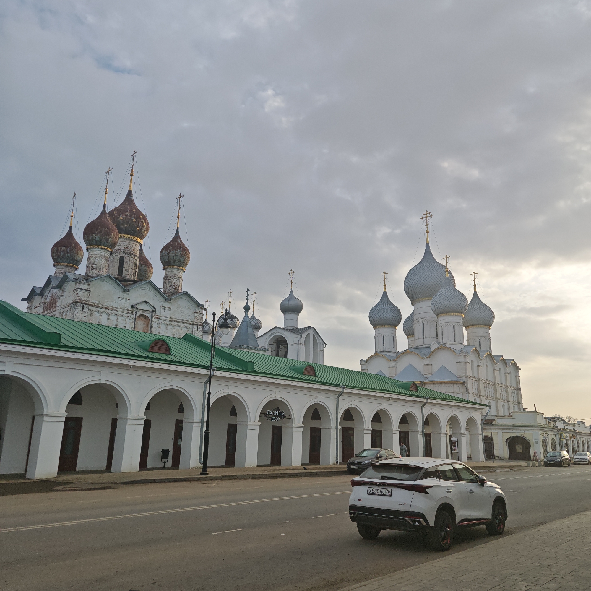 The image shows a street in Rostov, Russia, with a long white building with arches on the left side. Behind the white building are two Russian Orthodox churches. The church on the left has red onion domes, while the one on the right has silver domes. In front of the building is a street with a white car parked on the side. The sky is overcast with pale light filtering through. There are other cars parked further down the street. A sign on the building reads "ГОСТИНЫЙ ДВОР" which translates to "Trading Rows."