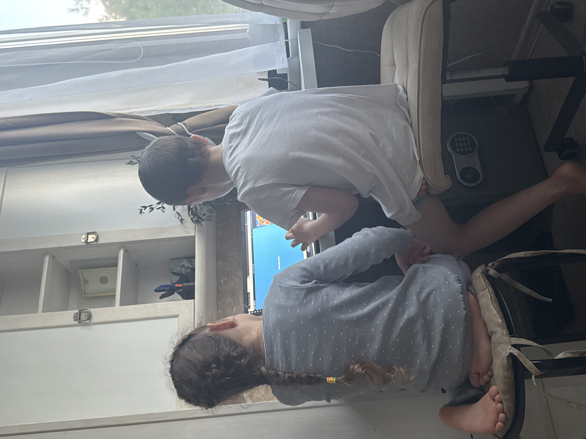A girl with braided hair and wearing a blue patterned shirt is sitting in front of a computer, next to a boy in a white t-shirt. The location is a room with a window and furniture, seemingly in a residential setting. The computer screen is visible, suggesting they may be engaged in an activity related to it. No information indicates this image was taken in Раздолье, Russia.