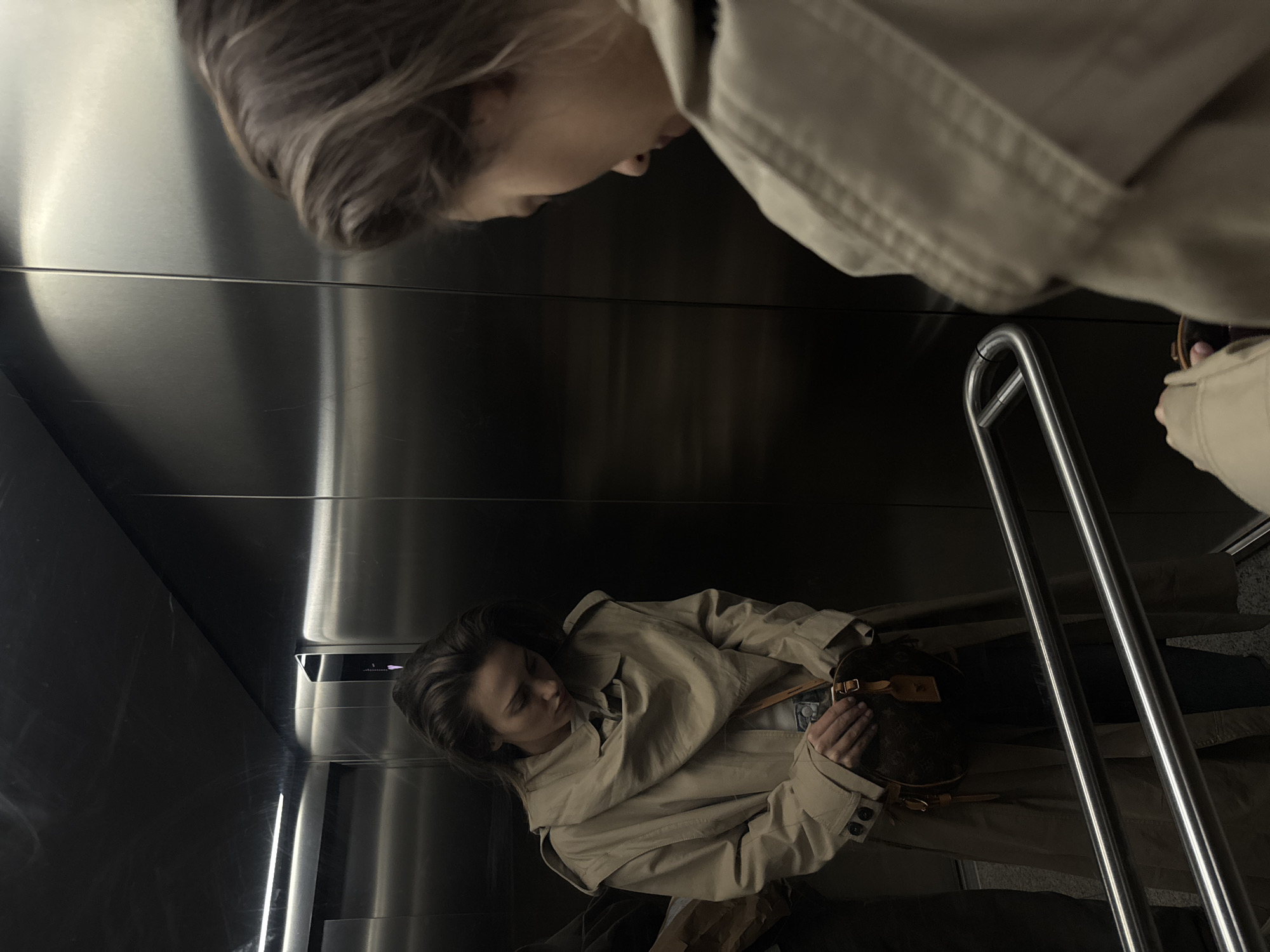 A mirrored scene shows a woman in a tan trench coat inside an elevator. The elevator has stainless steel walls and handrails. The woman's reflection appears to be looking down at her, with the actual woman in the lower half of the frame looking at a purse in her hands.