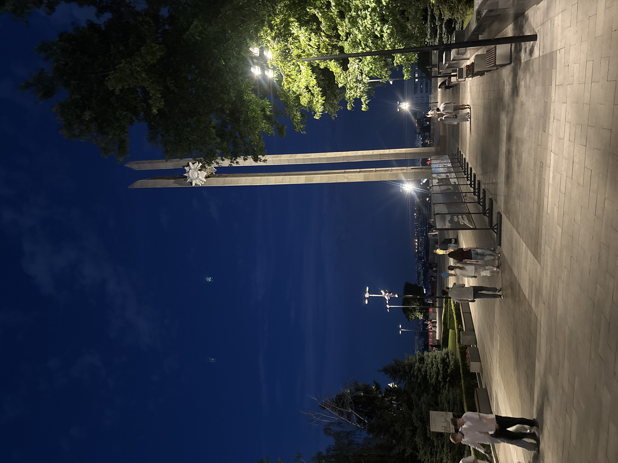 The image depicts a nighttime scene in Novaya Usman', Russia, likely a public square or promenade. Two prominent obelisk-like structures, possibly monuments, dominate the left side of the frame, silhouetted against a deep blue night sky. One obelisk features a decorative, star-like emblem. Trees with lush green foliage are visible, some illuminated by overhead lights, suggesting a park-like setting.
A paved walkway, made of rectangular stone tiles, stretches into the distance on the right side of the image. Several people are walking along this path, their figures casting shadows under the warm glow of streetlights. A few individuals are gathered near benches, engaged in conversation. Further down the walkway, a group of people appear to be looking at displays, perhaps an outdoor exhibition.
The time of day is clearly night, indicated by the dark sky and artificial lighting. The weather appears to be pleasant, with people dressed in casual summer clothing. The overall mood is serene and relaxed, with a sense of calm as people stroll through the illuminated public space. The city skyline is faintly visible in the background, suggesting an urban or suburban environment. No visible text is discernible in the image.