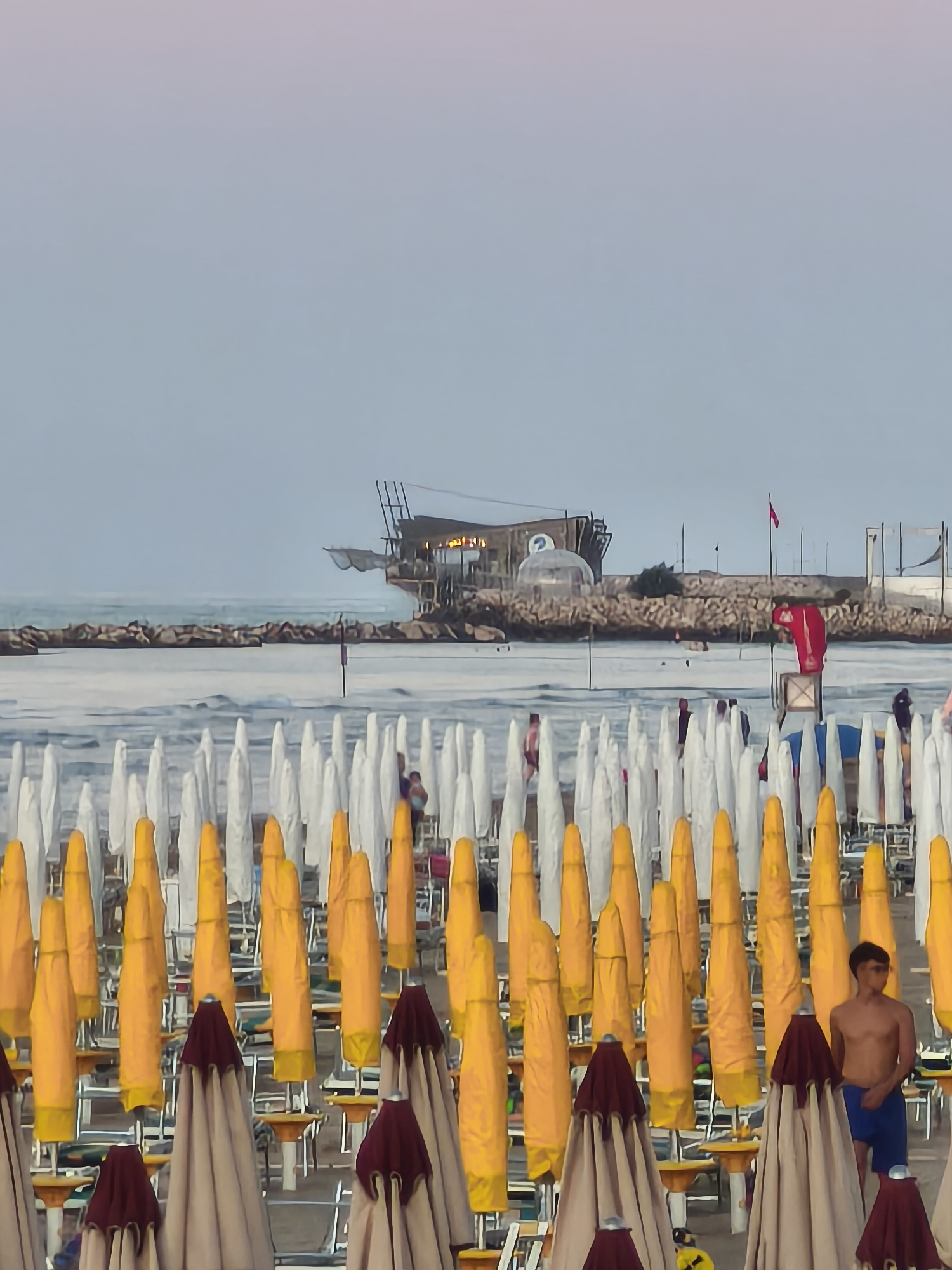 In San Salvo, Italy, a beach scene is depicted under a pale, hazy sky with hints of pink and purple at the horizon. Rows of closed beach umbrellas are lined up on the sand, predominantly yellow and white, with some beige and dark red umbrellas in the foreground. The umbrellas are organized in neat rows, suggesting a resort or public beach setting. In the middle ground, a man is visible standing between two umbrellas, wearing blue shorts. Further back, in the sea, a structure resembling a fishing platform or restaurant is built on stilts above the water, with additional structures and a dome-like enclosure visible on a rocky outcrop. Several people are seen on the beach and near the water, though their details are indistinct. The sea appears calm, with a rocky breakwater extending into the water. The overall atmosphere is that of a quiet, late afternoon or early evening on the coast.