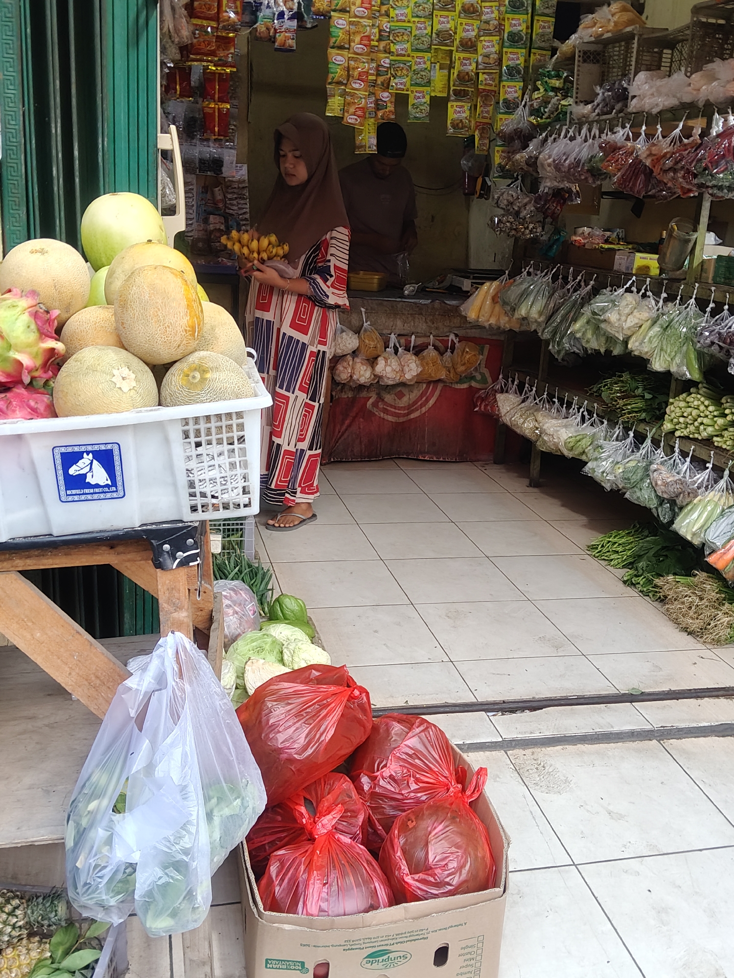 A street vendor stall in Mekarmukti, Indonesia, displays a variety of fresh produce and packaged goods. A young woman wearing a hijab and a patterned dress stands behind a table laden with melons, dragon fruit, and bananas. In the background, shelves are stocked with colorful bags of spices and dried goods, along with hanging bags of vegetables. Cabbages, green peppers, and other produce are visible in plastic bags and a cardboard box on the ground in the foreground. The stall appears to be open for business, with customers potentially browsing the offerings.