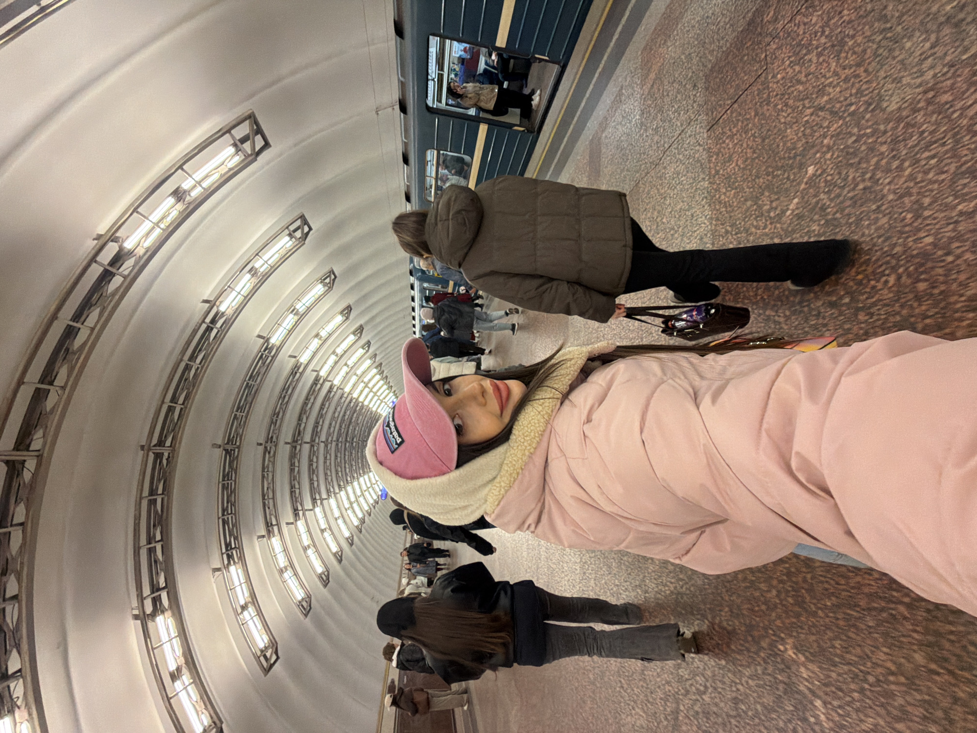 The image shows a subway station in Saint Petersburg, Russia. The station features a curved, arched ceiling with rows of lights. Several people are standing on the platform, near a subway train. One woman in the foreground is wearing a pink jacket and a pink cap. Another person is wearing a brown jacket and carrying a black bag. Others are standing or walking in the background.